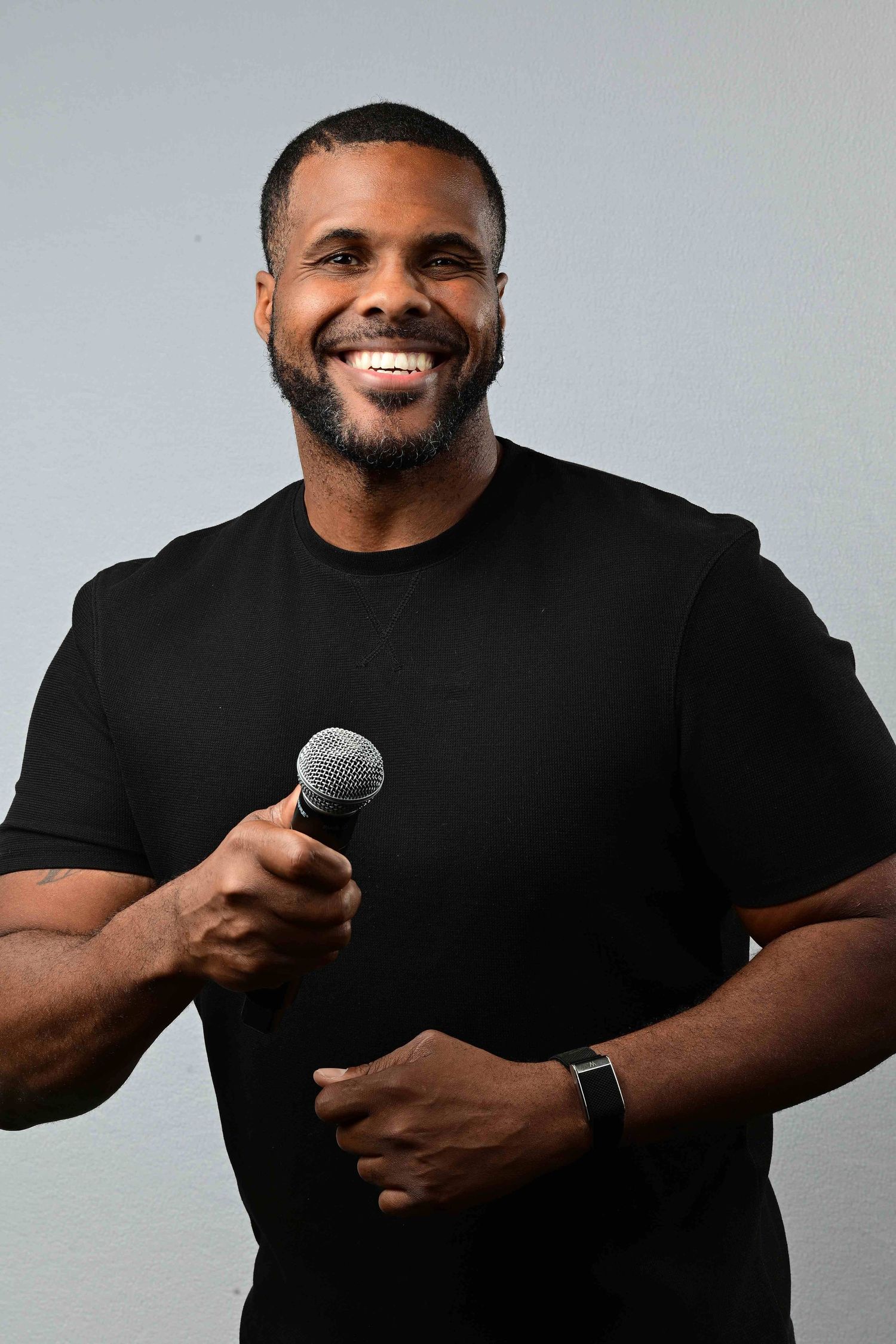 Smiling man with a short beard in a black t-shirt and smartwatch holds a microphone against a plain light gray background.