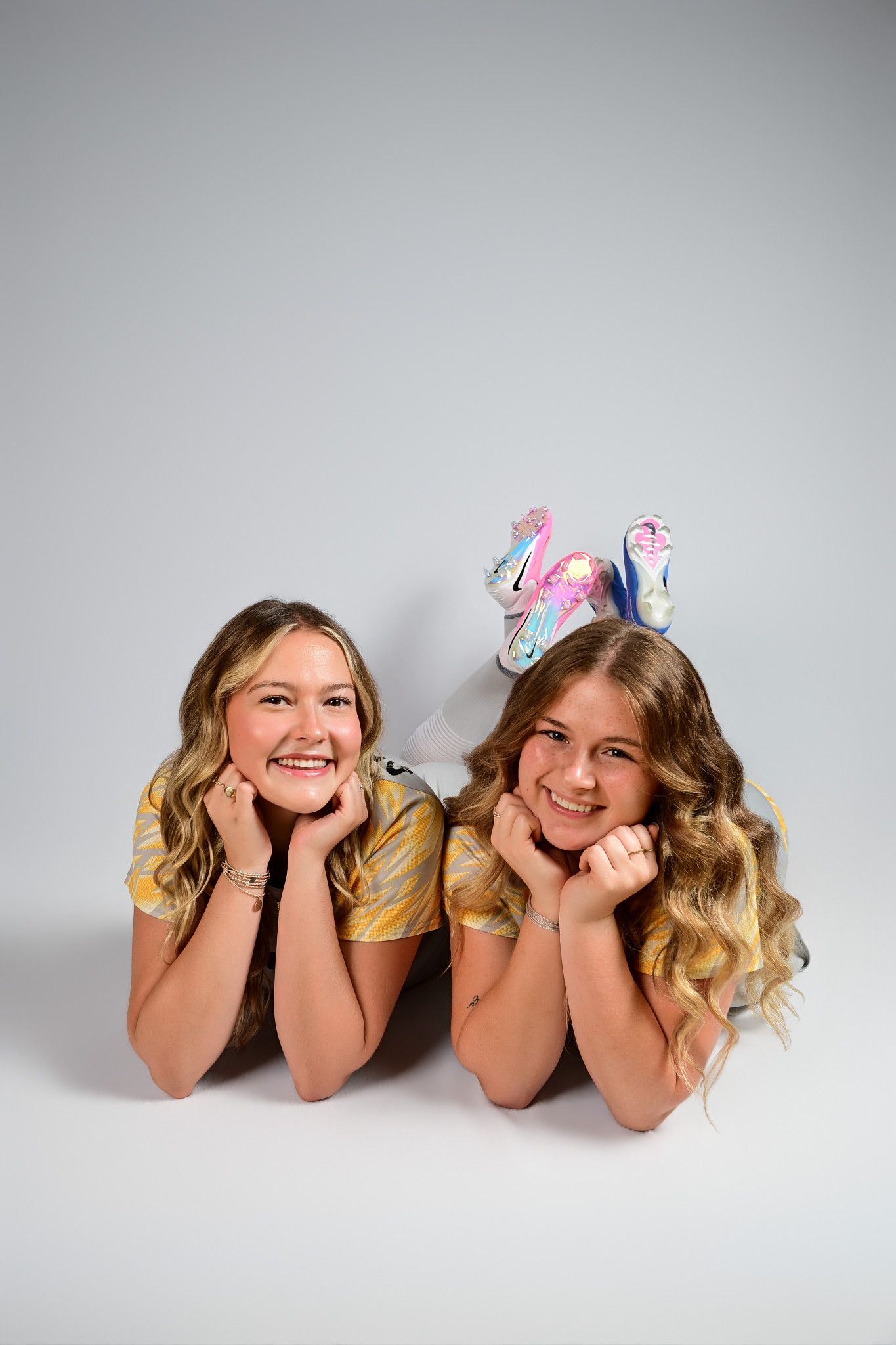 Two smiling teenage girls lying on floor, one wearing colorful roller skates, posing with hands on cheeks against gray background.