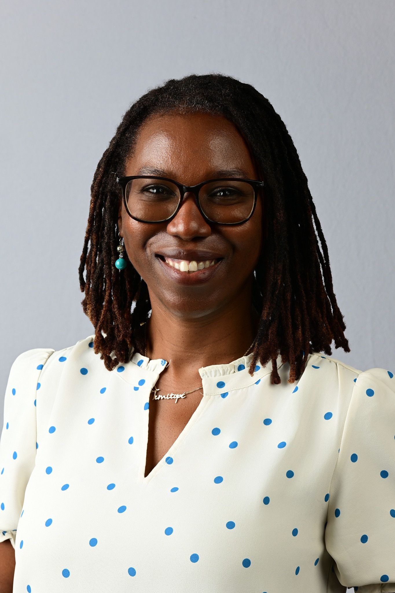 Smiling woman with dreadlocks and glasses wearing a white polka dot blouse, professional headshot portrait.