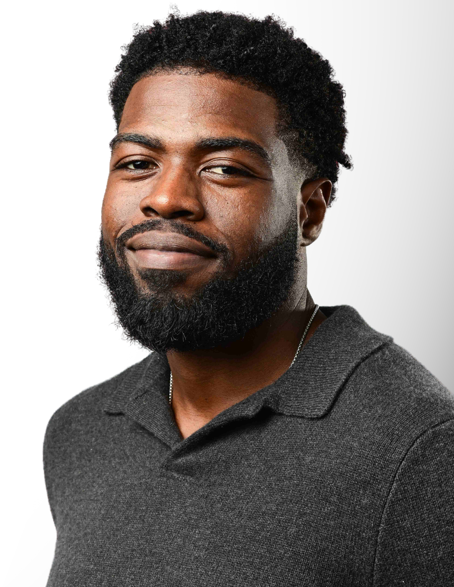 Young Black man with a beard and short curly hair wearing a dark gray polo shirt against a white background.