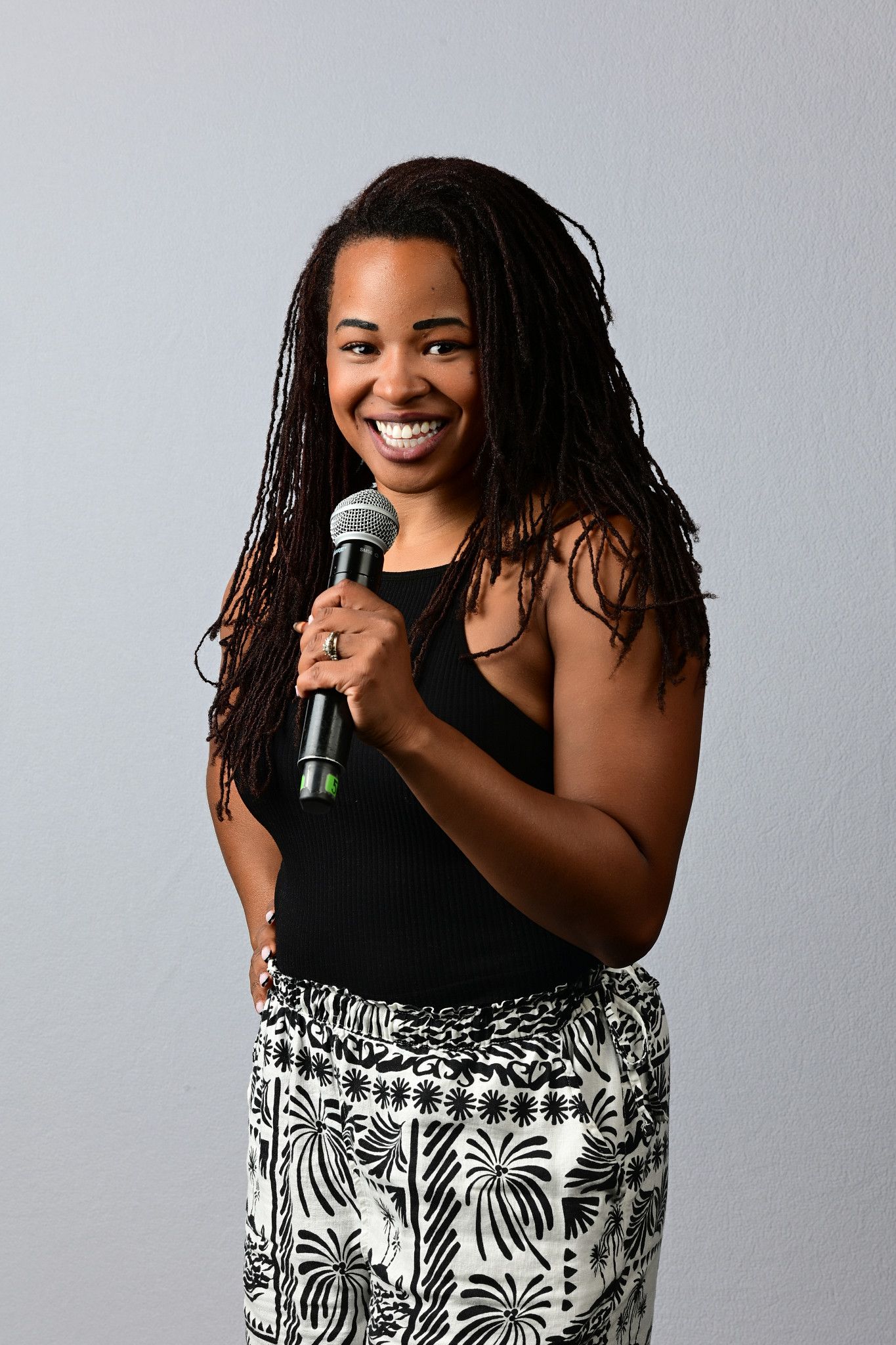 Smiling woman with long locs holds a microphone, wearing a black tank top and a black and white palm print skirt.