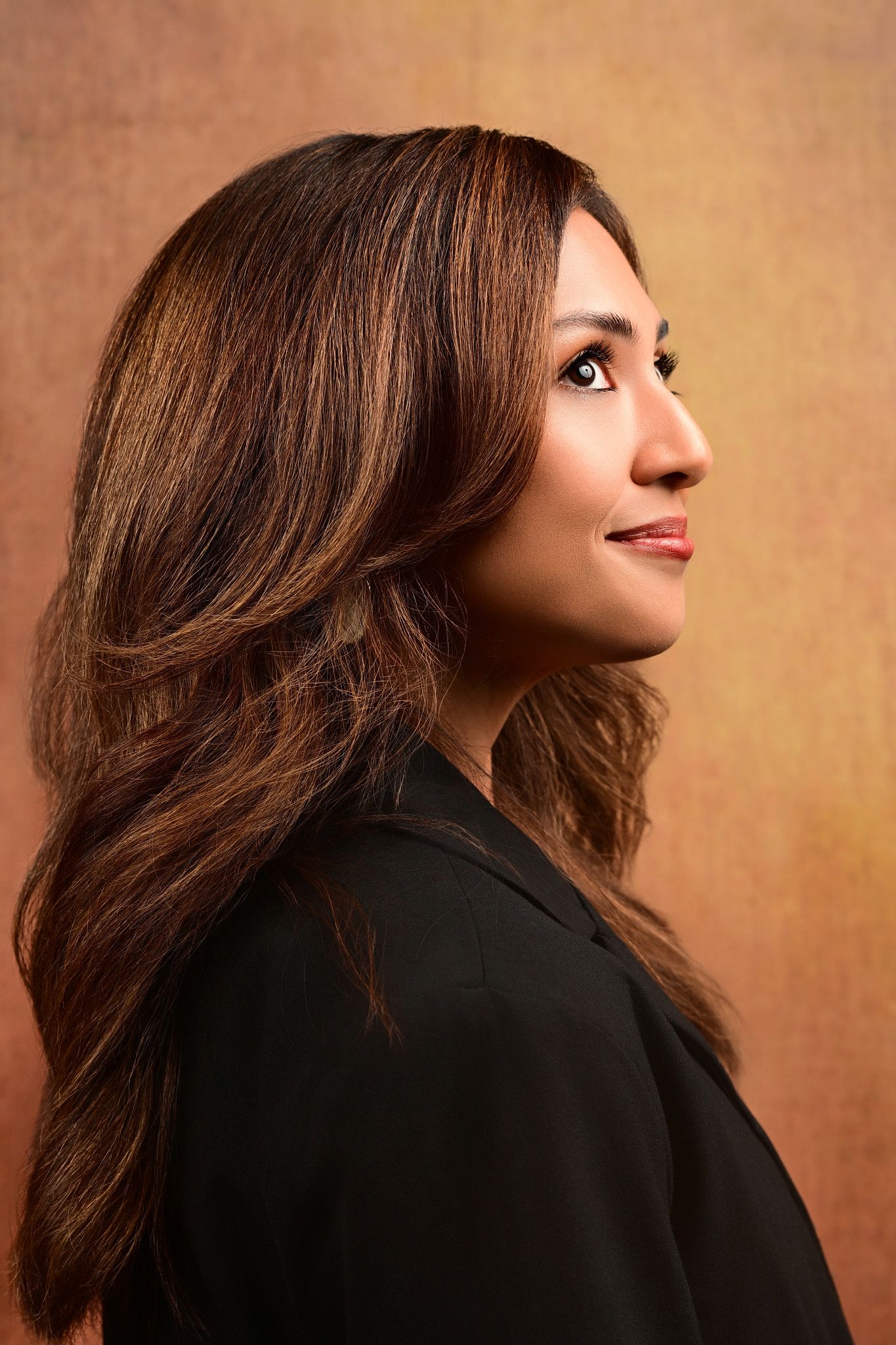Side profile portrait of a woman with long, wavy brown hair and a black blazer, gazing upward against a warm amber textured background.