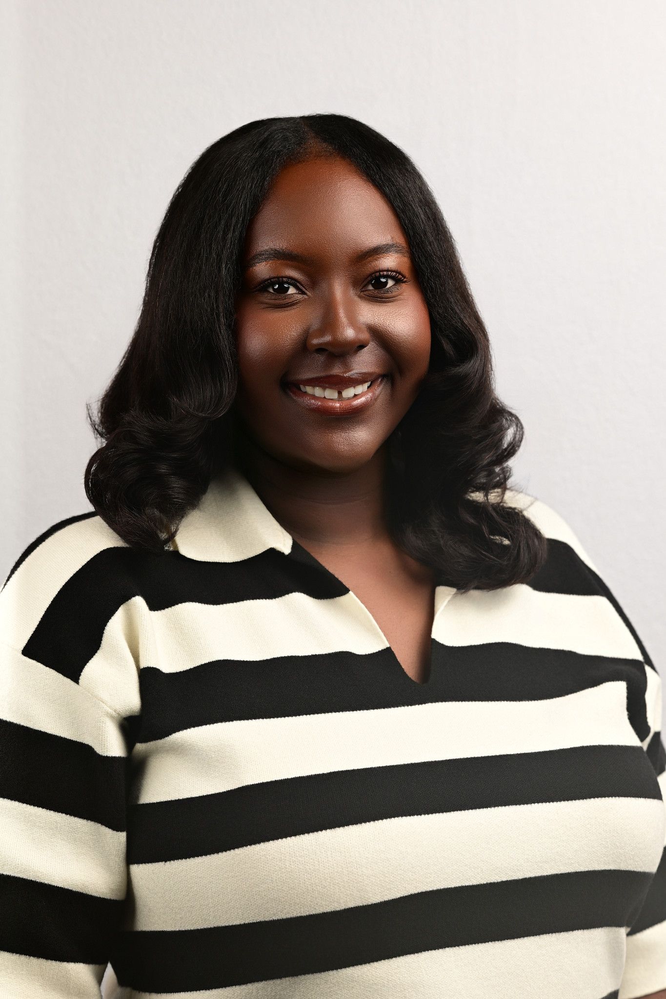 Smiling woman with shoulder-length wavy black hair wearing a black and cream striped collared sweater against a light gray background.