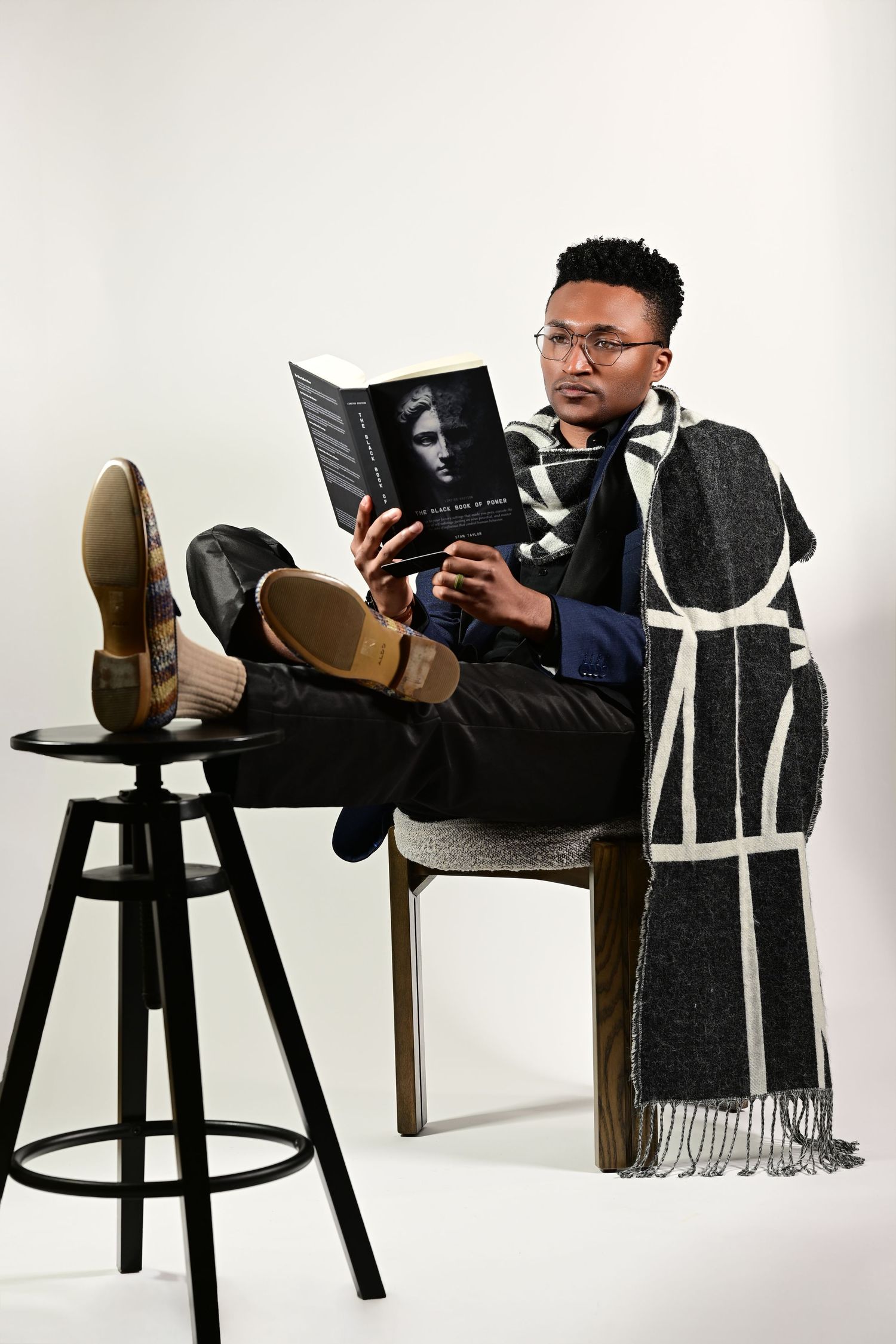 A stylish man reading a book while relaxing in a chair with feet up, wearing glasses and a plaid scarf.