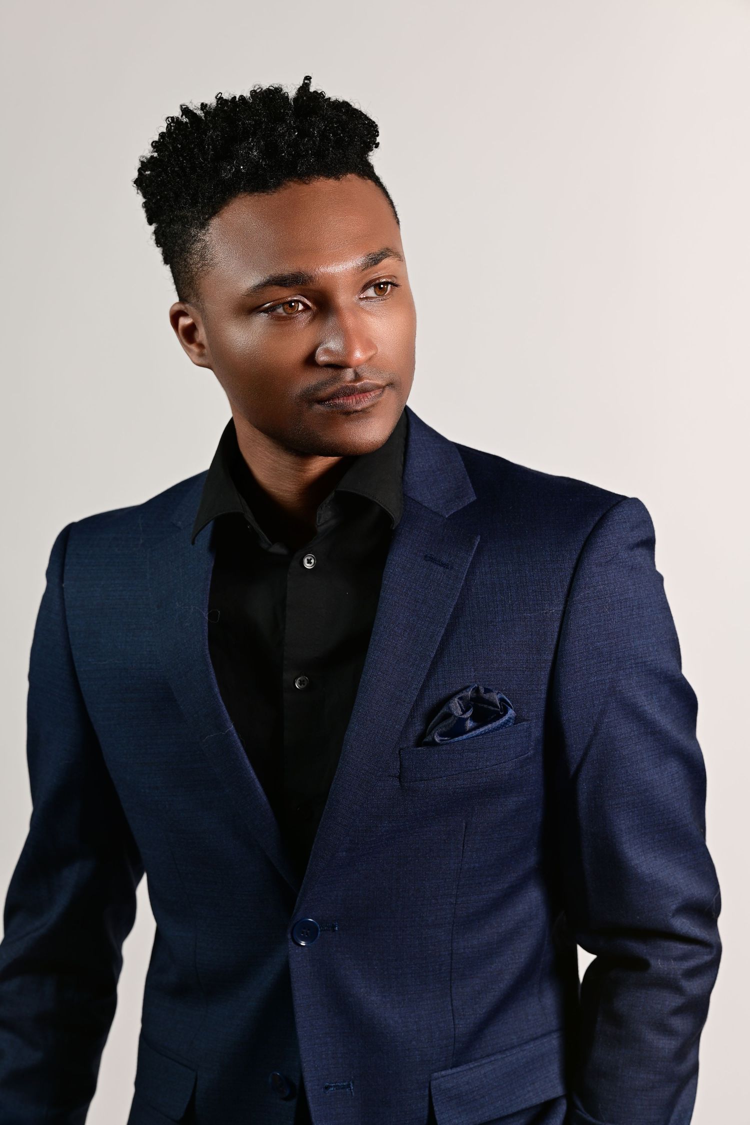 A young Black man in a navy blue suit and black shirt with pocket square poses confidently against a gray background.