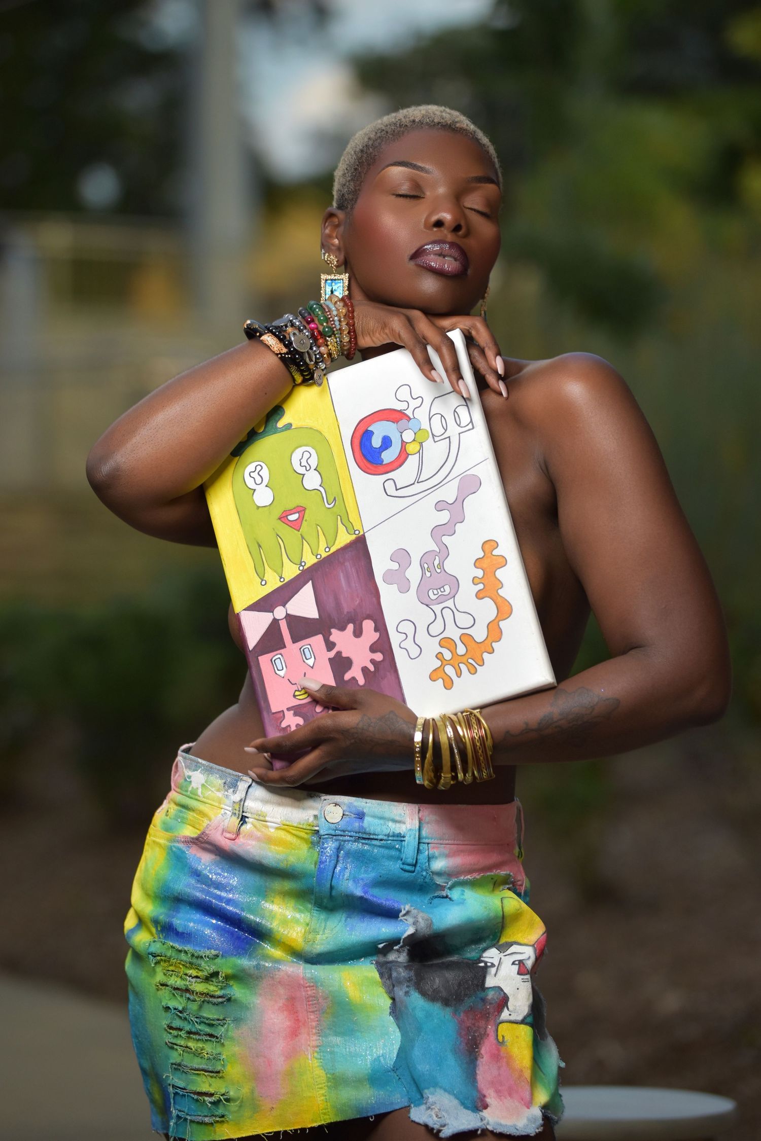 Stylish Black woman with cropped hair posing with vibrant painted canvas in tie-dye skirt outdoors.