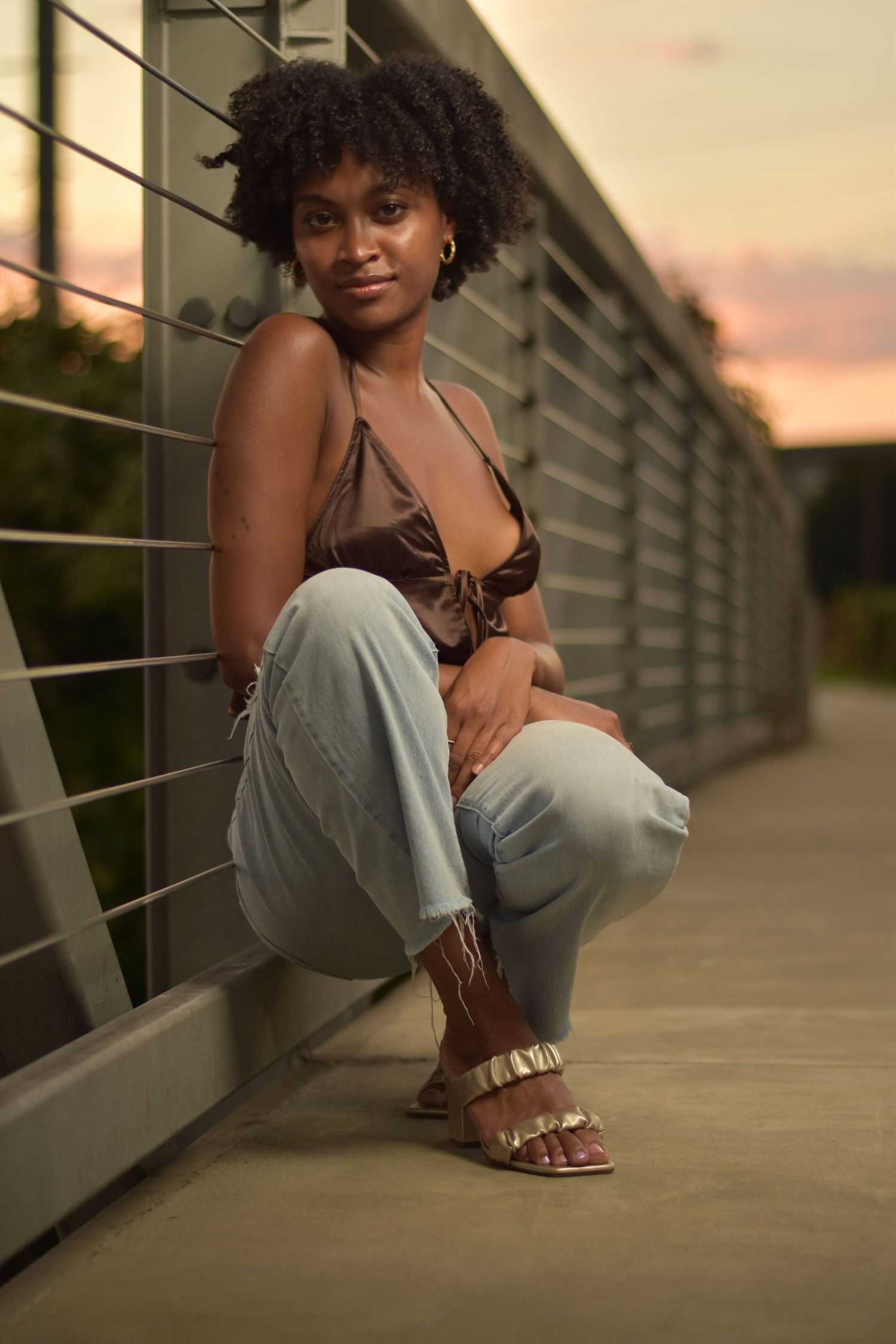 Young Black woman in brown satin crop top and light jeans crouching by metal railing at sunset.