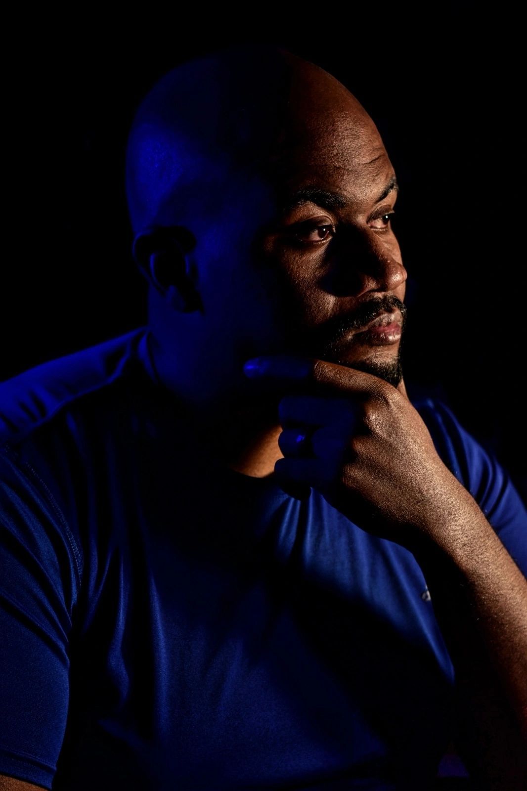 A bald Black man with a goatee poses thoughtfully with hand on chin, dramatically lit with blue light on black background.