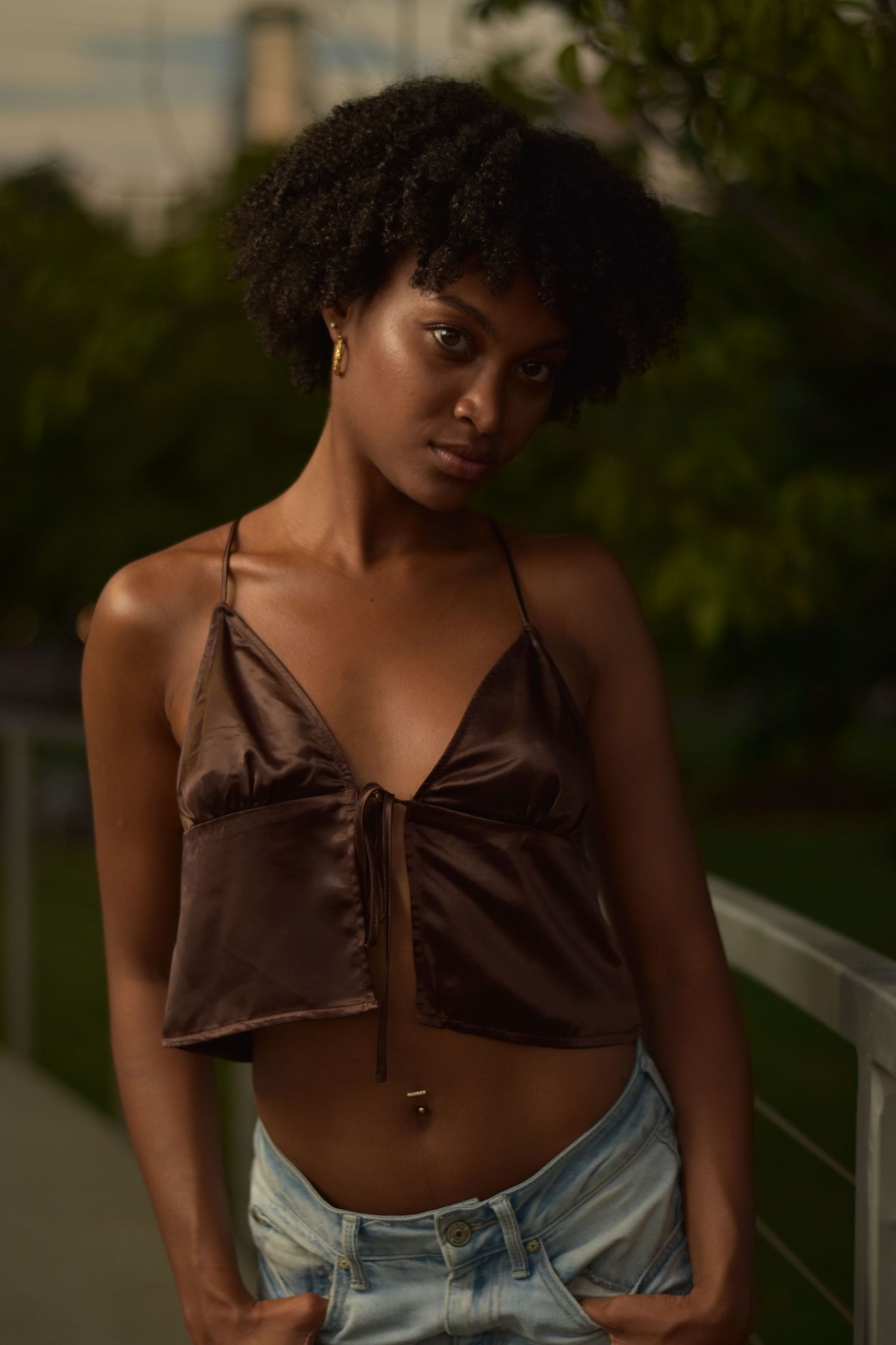 Black woman in brown satin halter crop top and light jeans standing by railing at sunset.