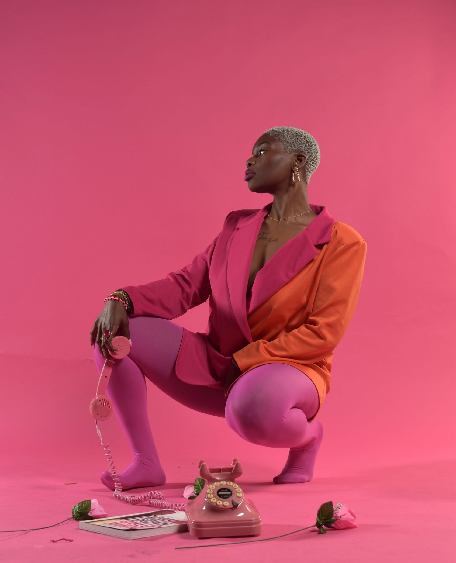 Fashionable Black woman in pink orange colorblock suit crouching beside vintage pink telephone on pink background.