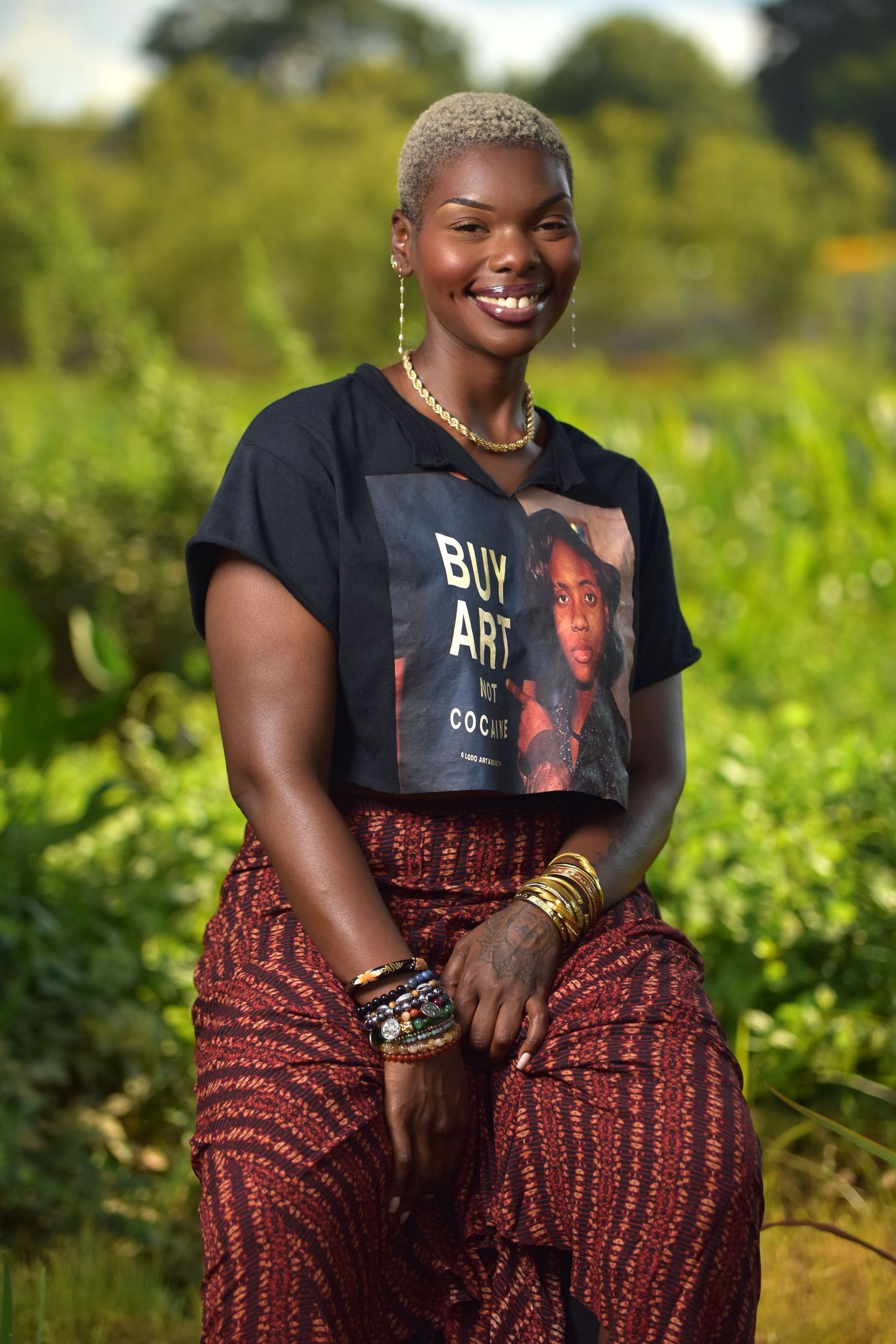 Smiling Black woman with short blonde hair wearing Buy Art graphic tee and red patterned pants in lush green setting.