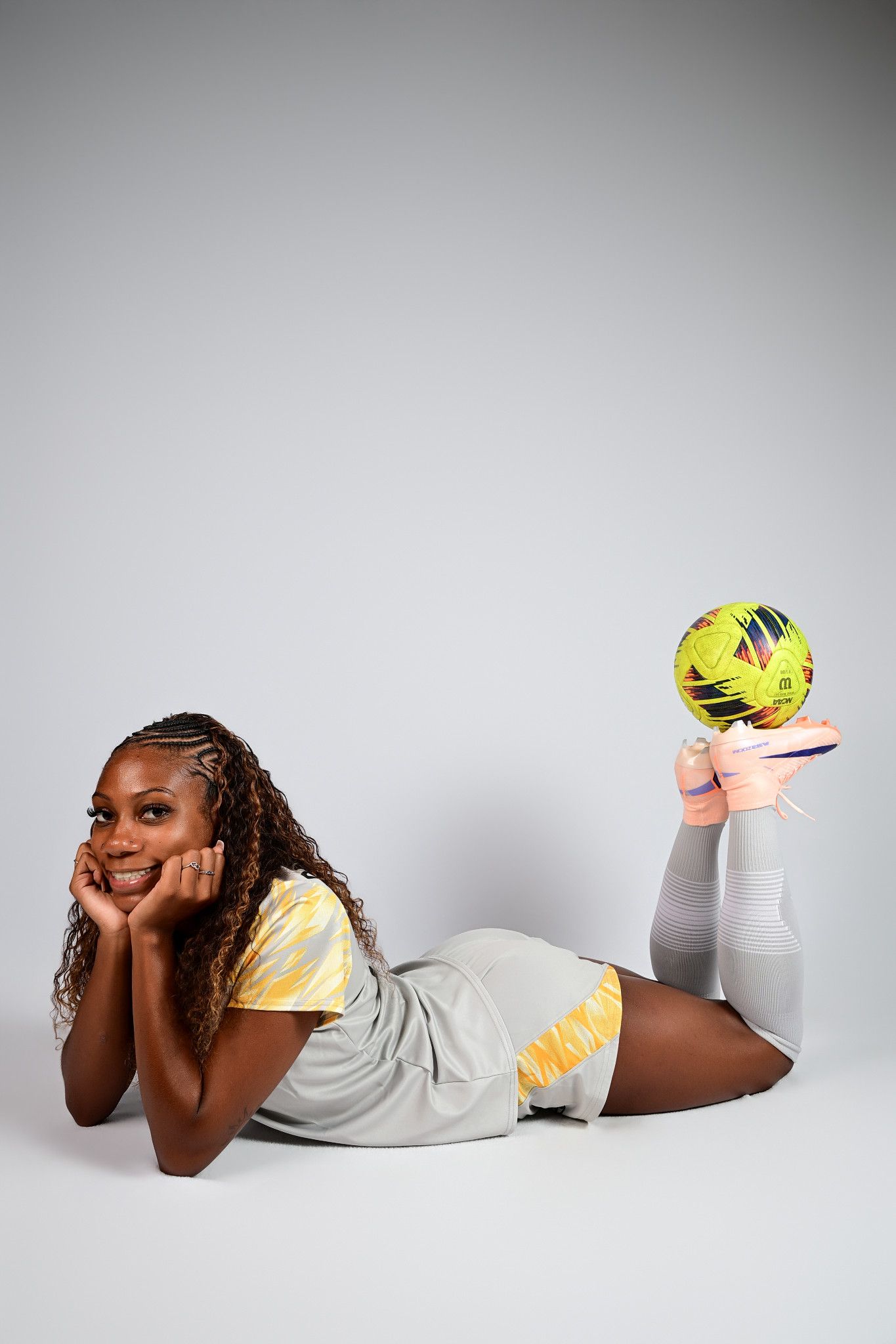 Young woman in athletic wear lying down smiling, balancing a yellow soccer ball on her foot.