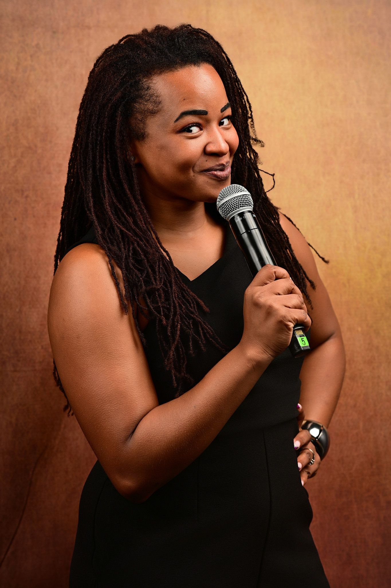 Woman with long locs in a black sleeveless dress holds a microphone and smiles playfully against a warm orange-brown backdrop.