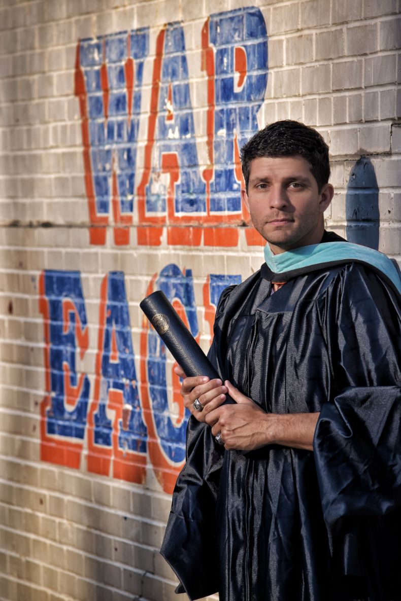 A male graduate in black cap and gown holds a diploma in front of a 'War Eagle' painted brick wall.