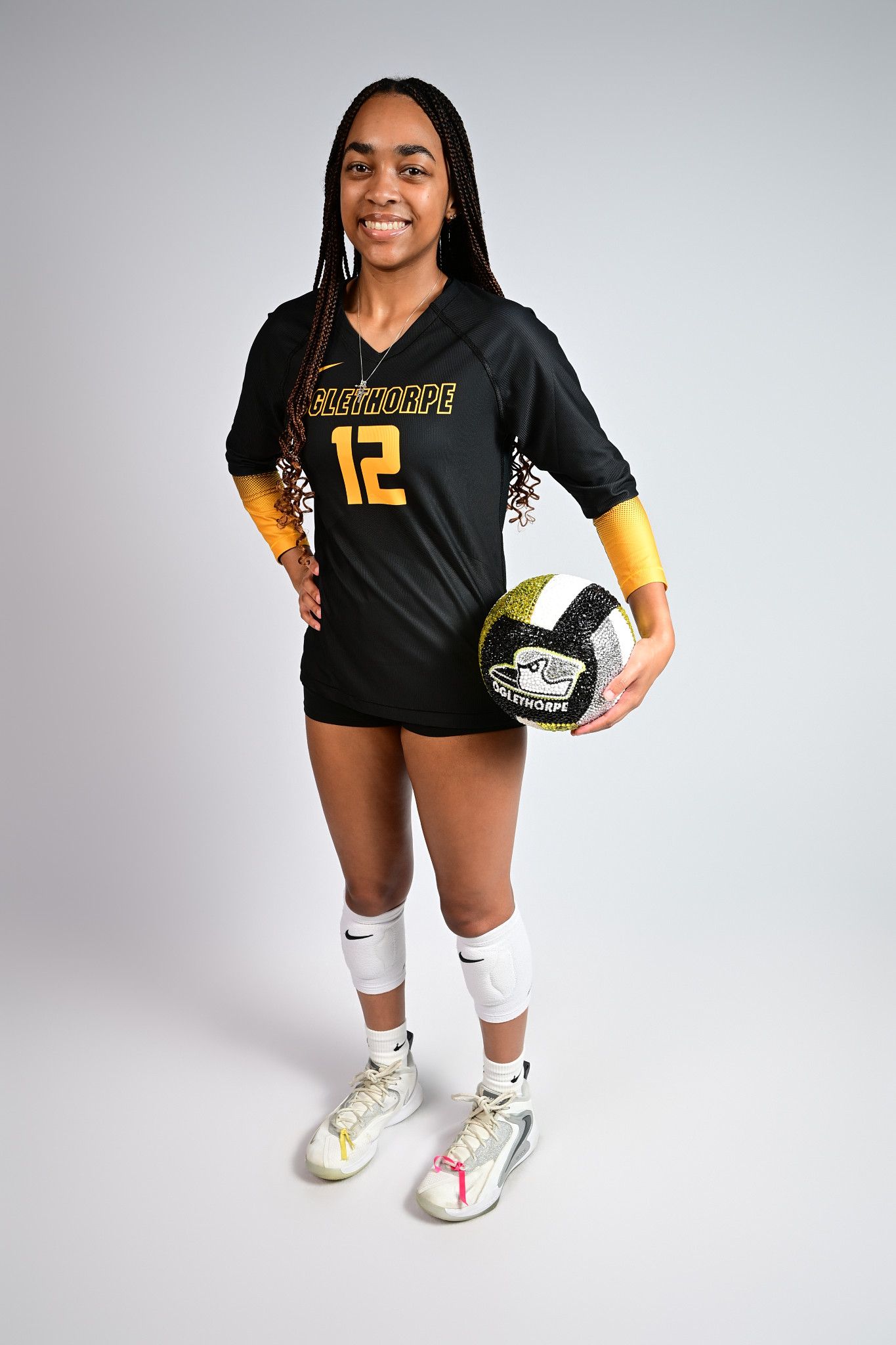 A smiling female volleyball player in a black number 12 jersey holds a ball during an official team photo shoot.