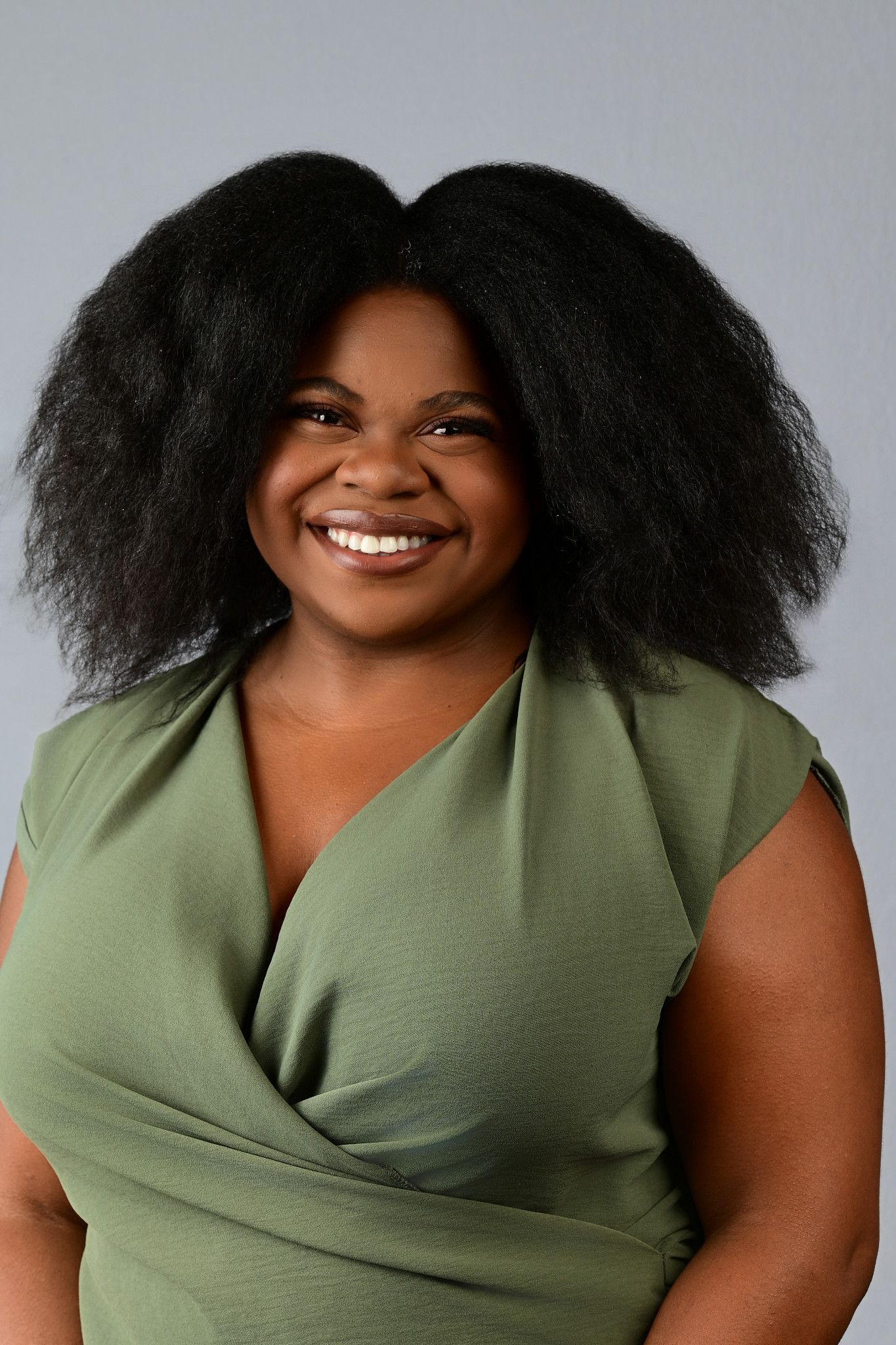 Smiling Black woman with natural hair wearing a green wrap top, posing for a professional headshot photo.