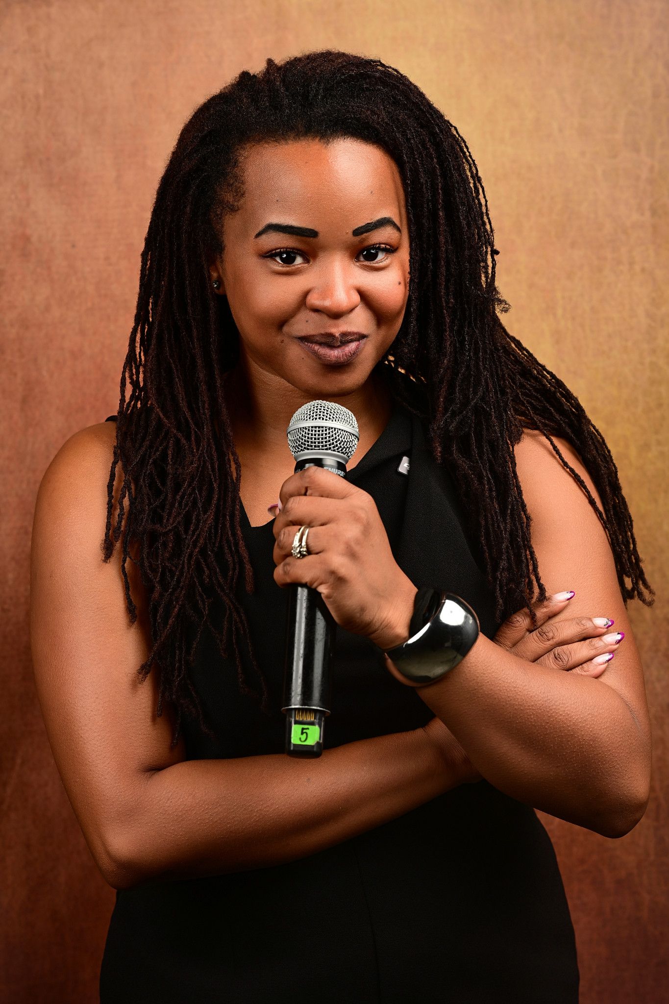 Smiling woman with long locs in a black sleeveless top holds a microphone with arms crossed against a warm bronze backdrop.