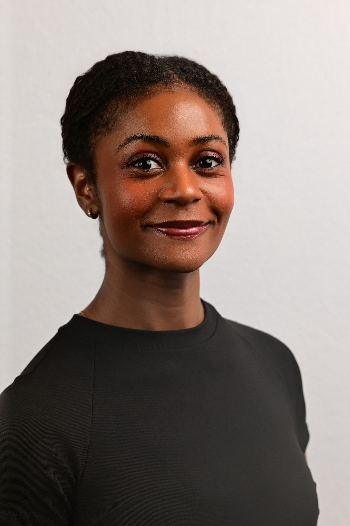 Smiling woman with braided updo and stud earrings wearing a black crew-neck top against a plain light gray studio background.