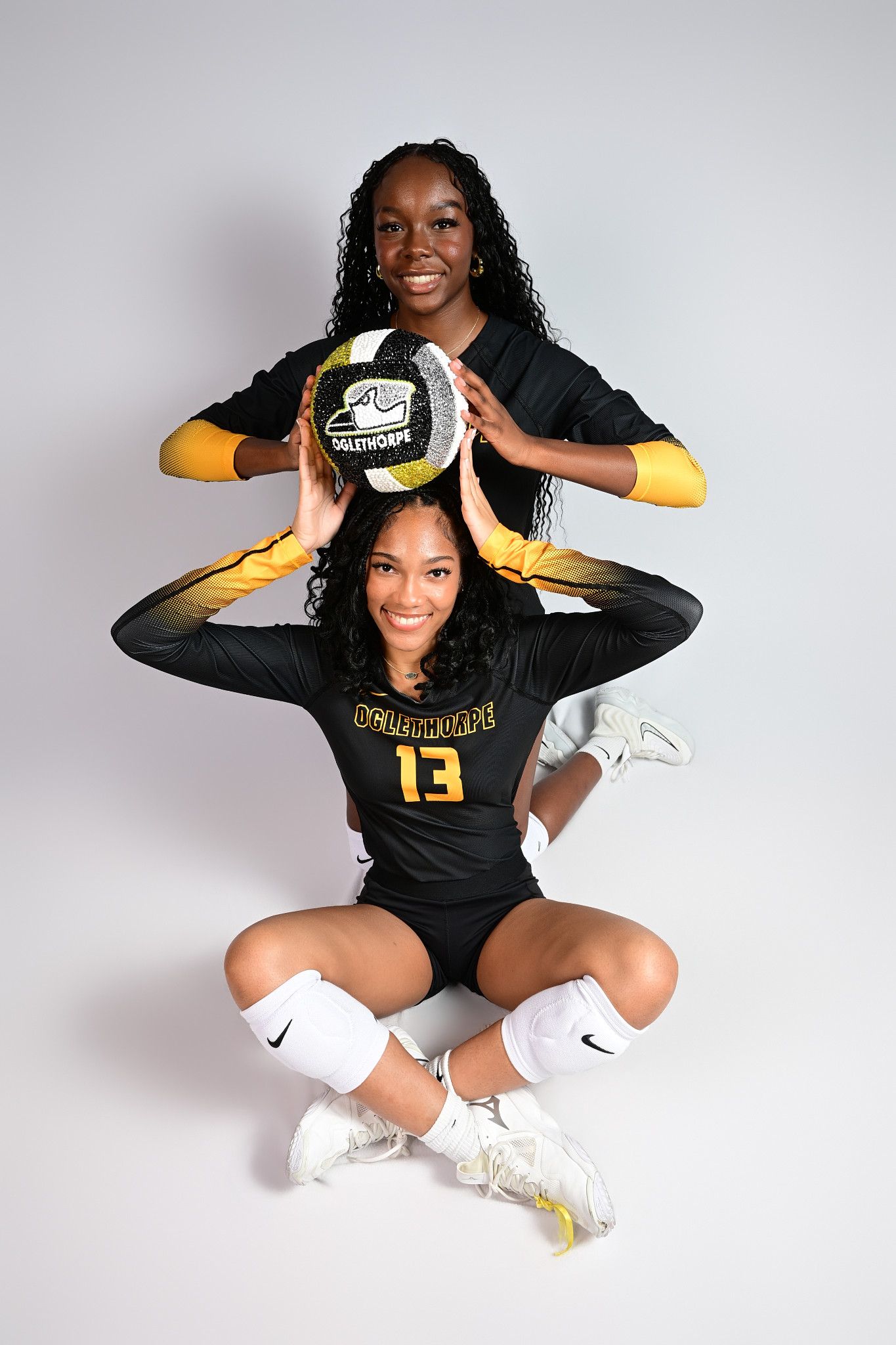 Two female volleyball players in black and gold uniforms pose together, one seated holding a volleyball, for a team portrait.