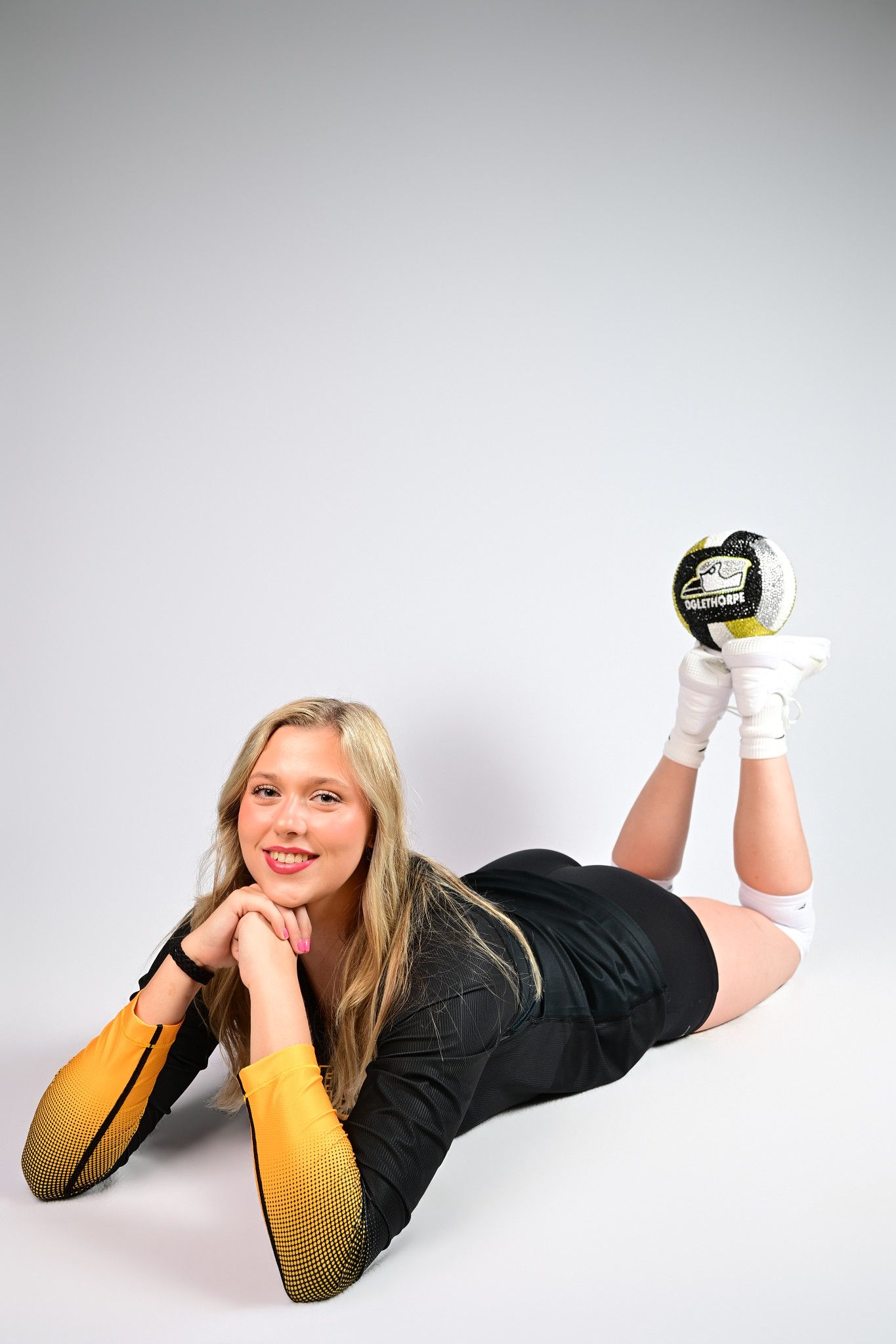 Young blonde woman in black sports uniform lying down holding a soccer ball wearing goalkeeper gloves.