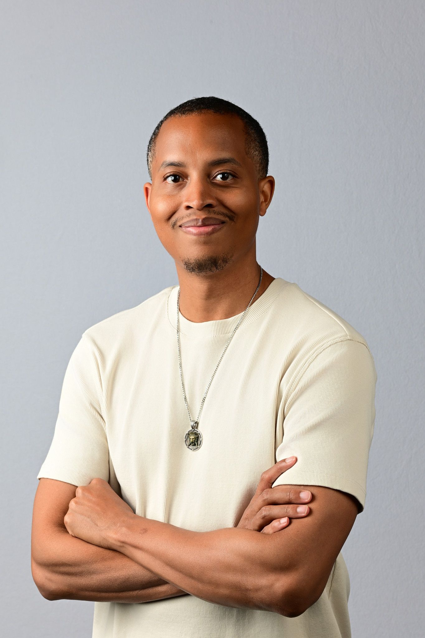 Young Black man in cream t-shirt with silver pendant necklace, arms crossed, smiling confidently against gray background.