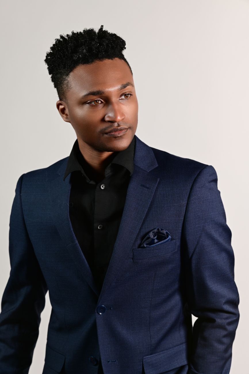 A young Black man in a navy blue suit with black shirt and pocket square poses confidently against a light gray background.