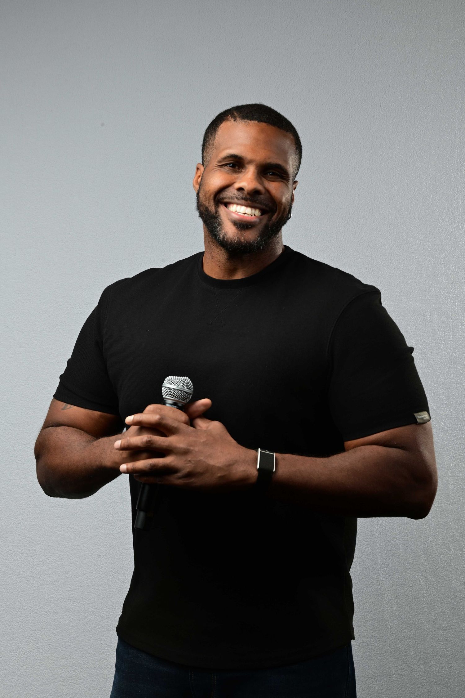 Smiling bearded man in a black t-shirt and smartwatch holds a microphone with clasped hands against a plain gray background.