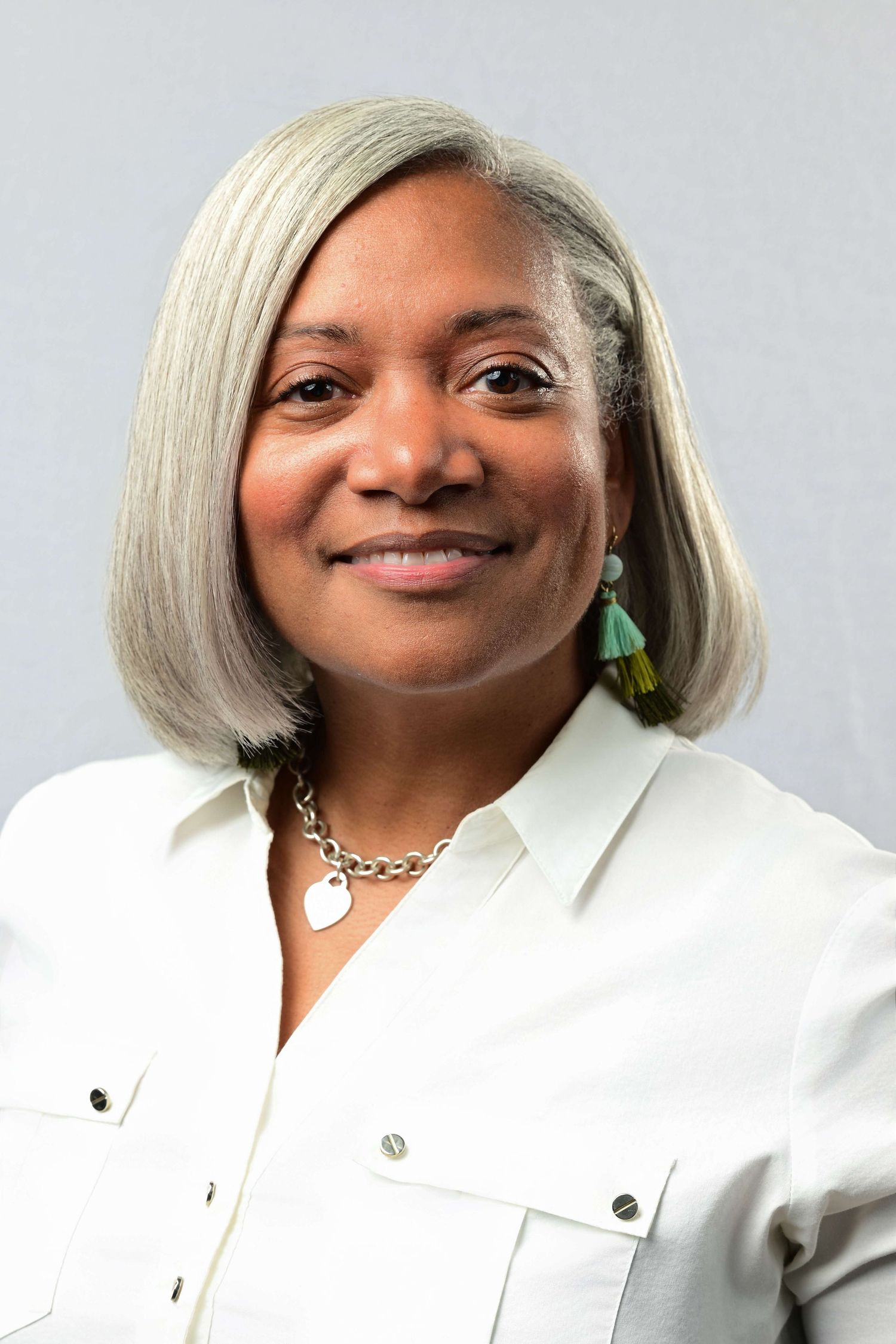 Professional headshot of a woman with gray bob hair, teal earrings, silver necklace, and white button-up shirt.