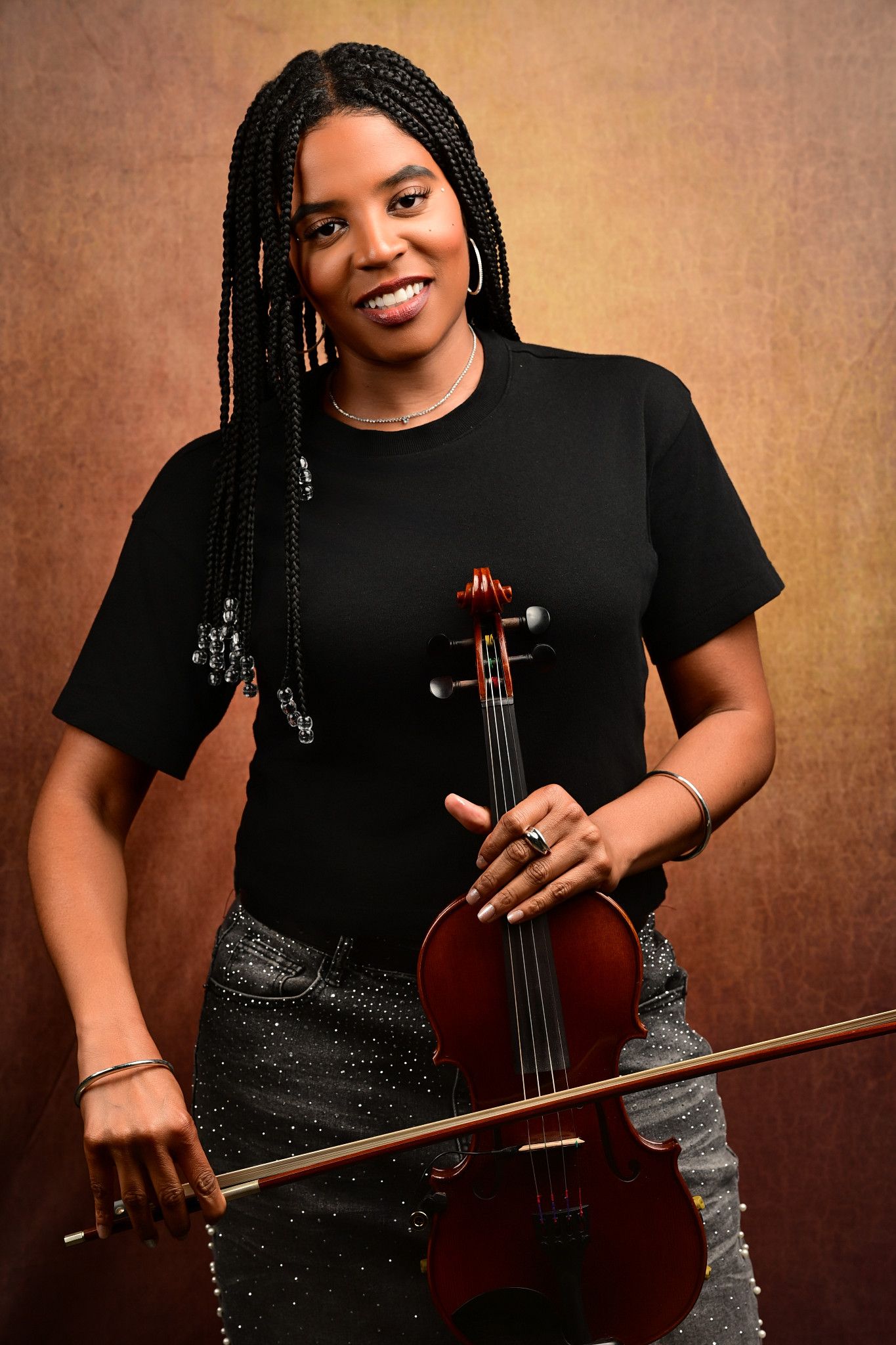 Smiling woman with long braids in a black top and studded skirt holds a violin and bow against a warm, textured brown studio backdrop.