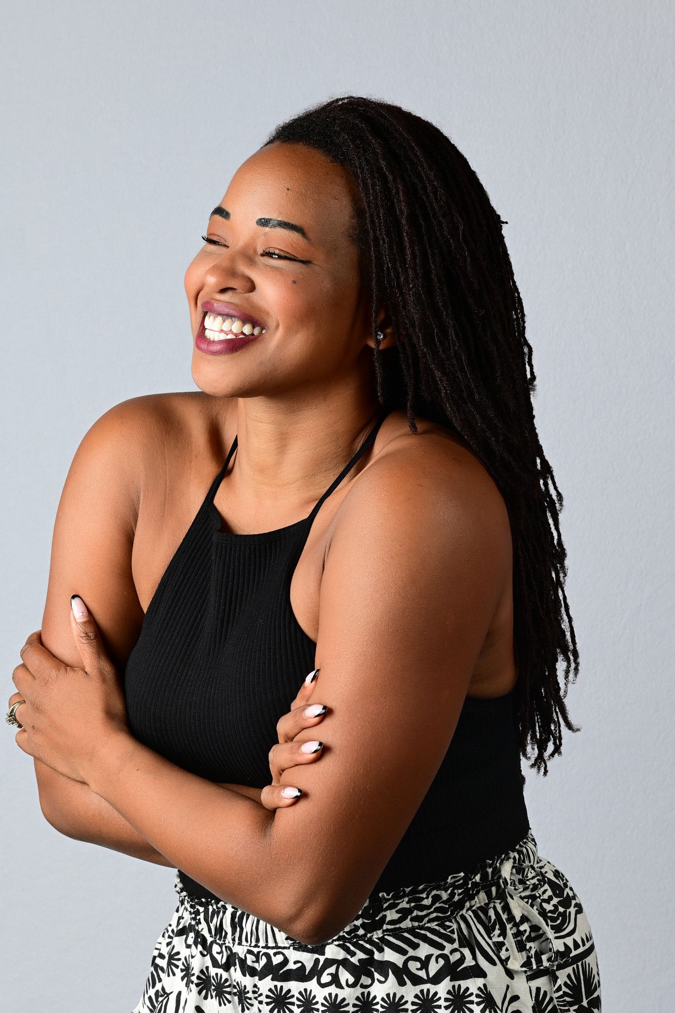 A joyful woman with long locs and a black halter top laughs with her arms crossed against a soft gray studio background.