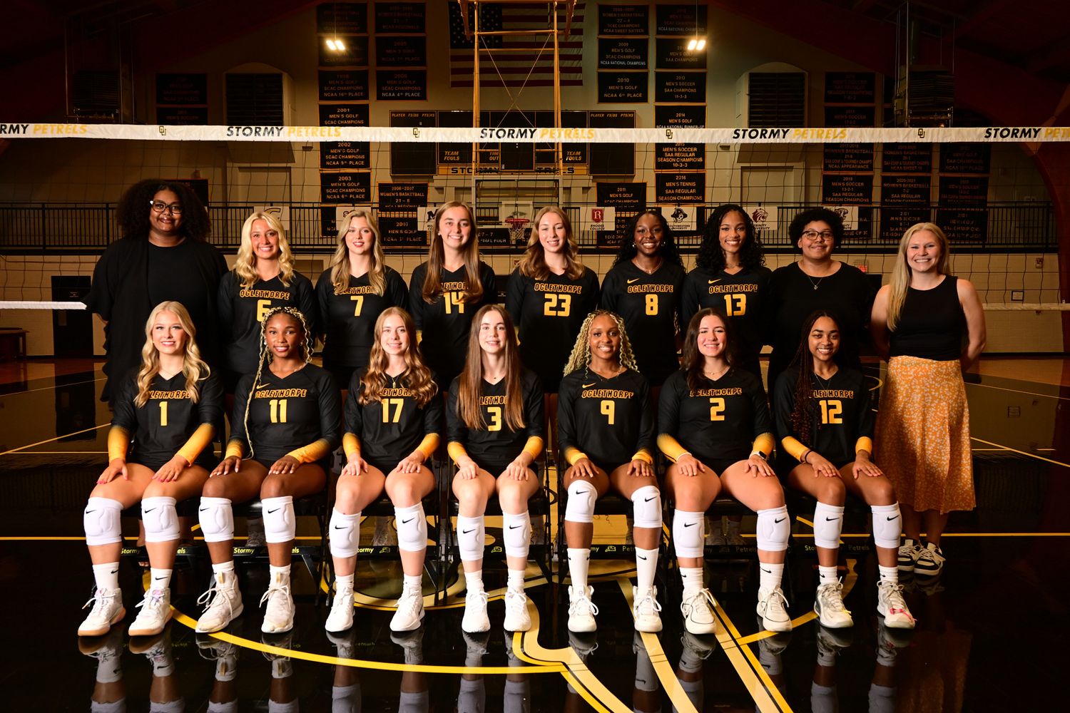 Women's volleyball team poses in black and gold uniforms for official team photo on indoor court.
