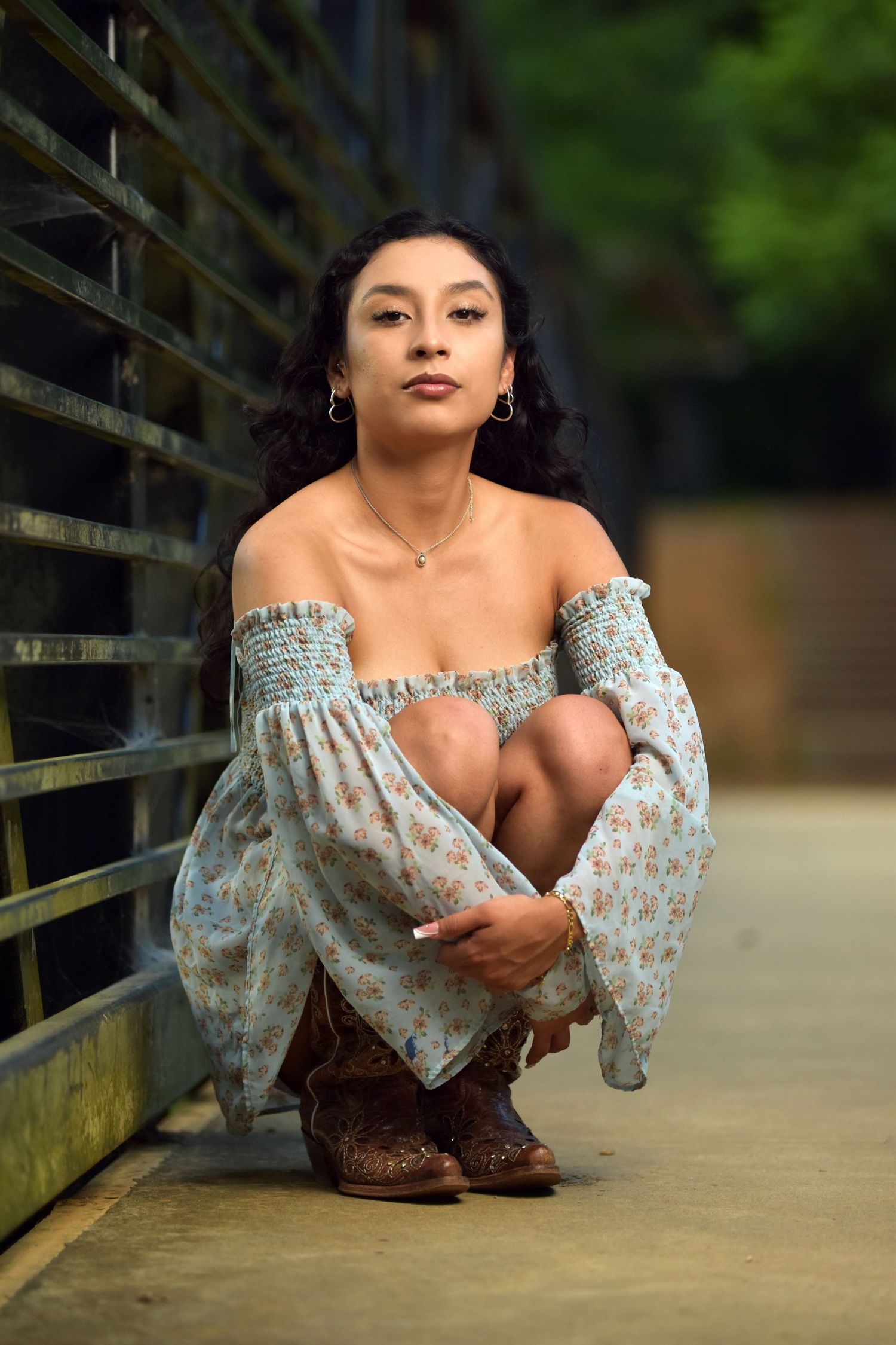 Young woman in floral off-shoulder dress and cowboy boots crouching against rustic metal fence.