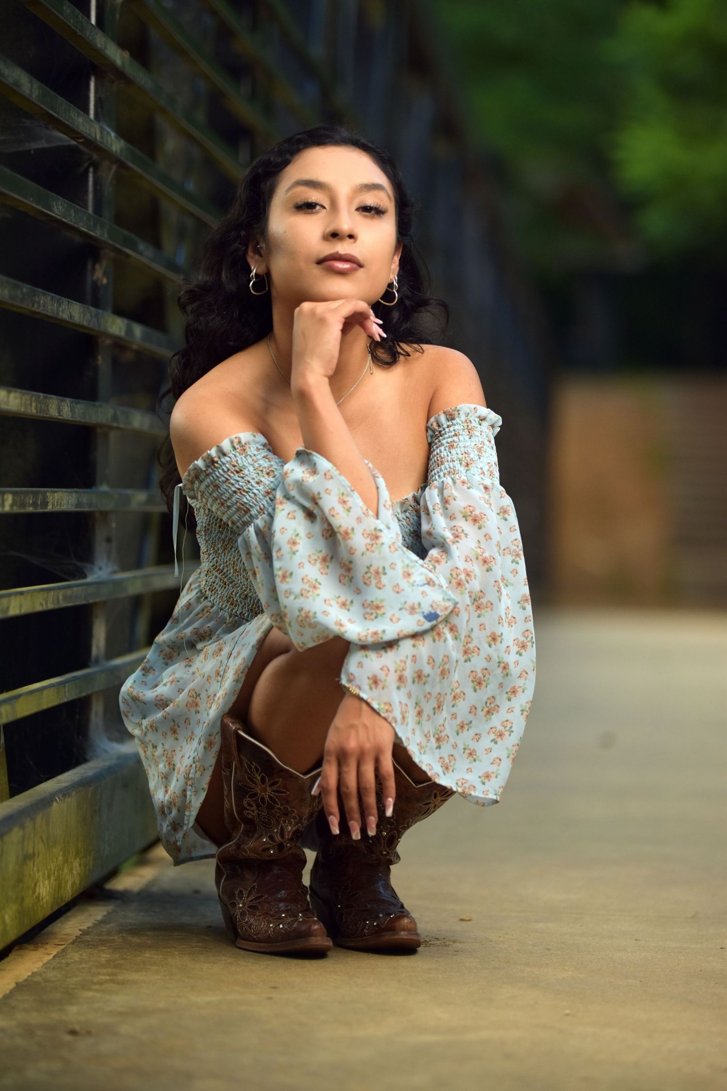 Young woman in floral off-shoulder dress and brown cowboy boots posing crouched by metal railing.