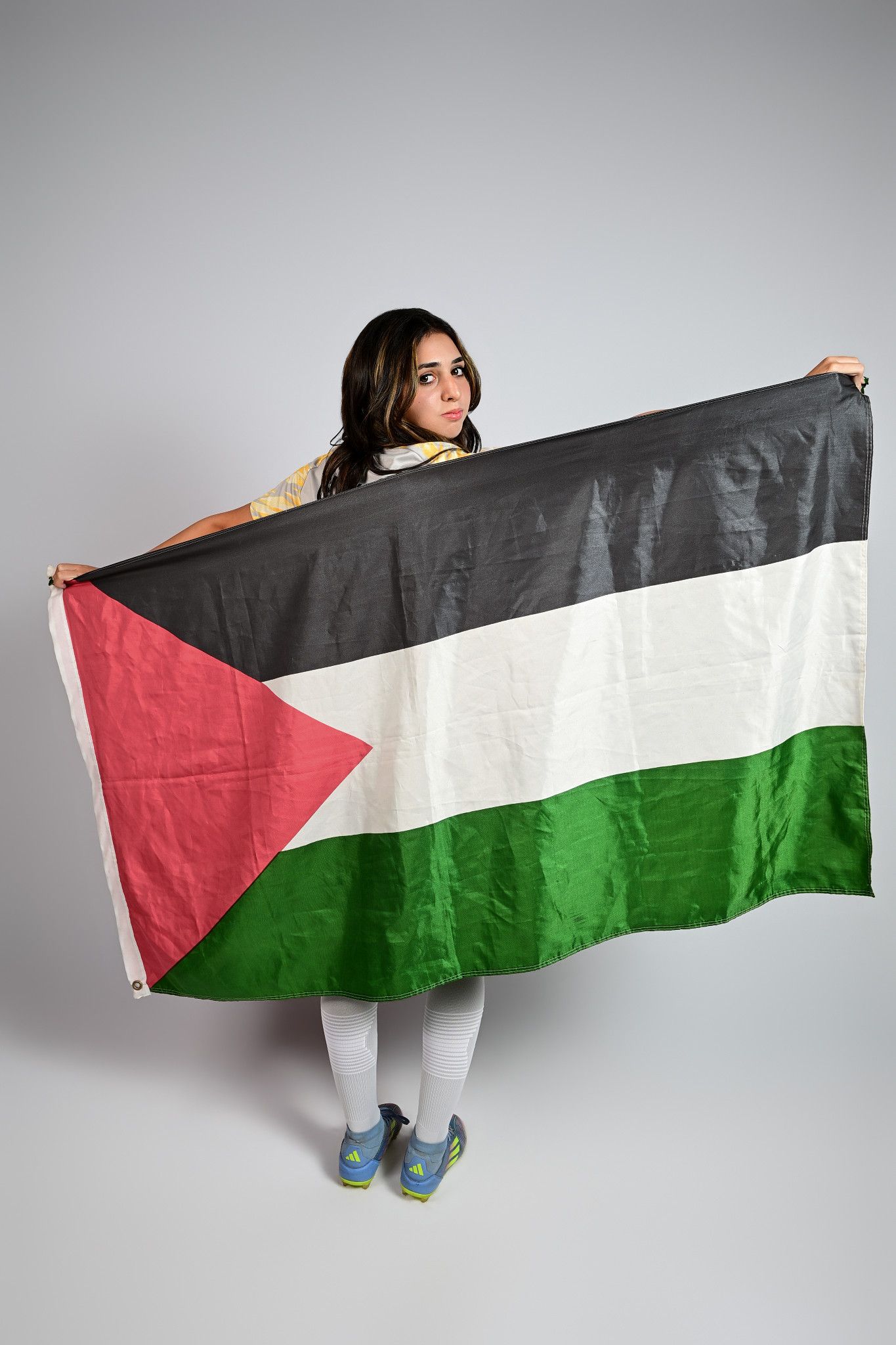 Young woman holding a large Palestinian flag in front of her body against a gray background.