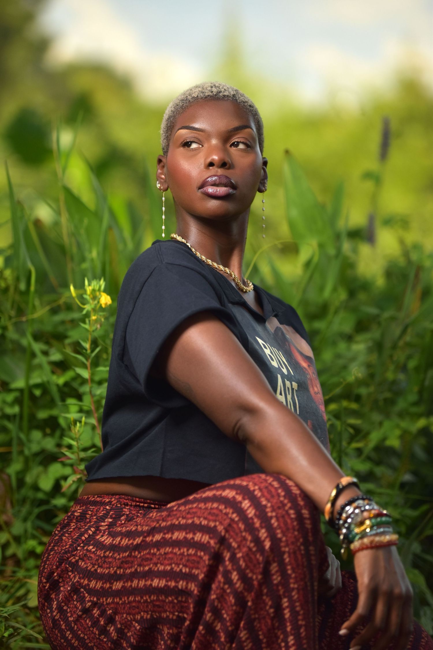 Young Black woman with blonde cropped hair sitting among green plants, wearing black graphic tee and red patterned skirt.