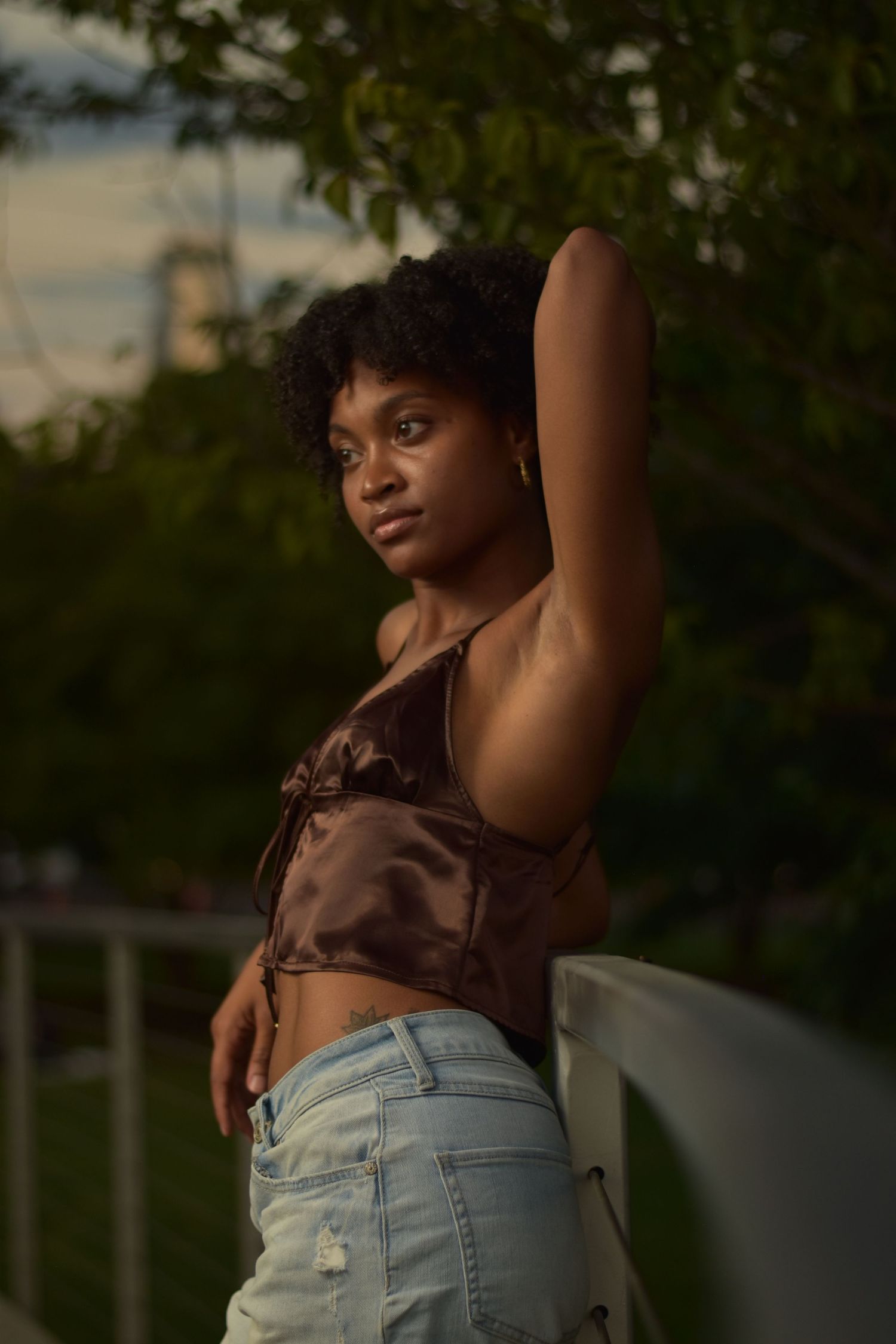 Black woman in brown satin crop top raising arm while leaning on railing with green trees behind.