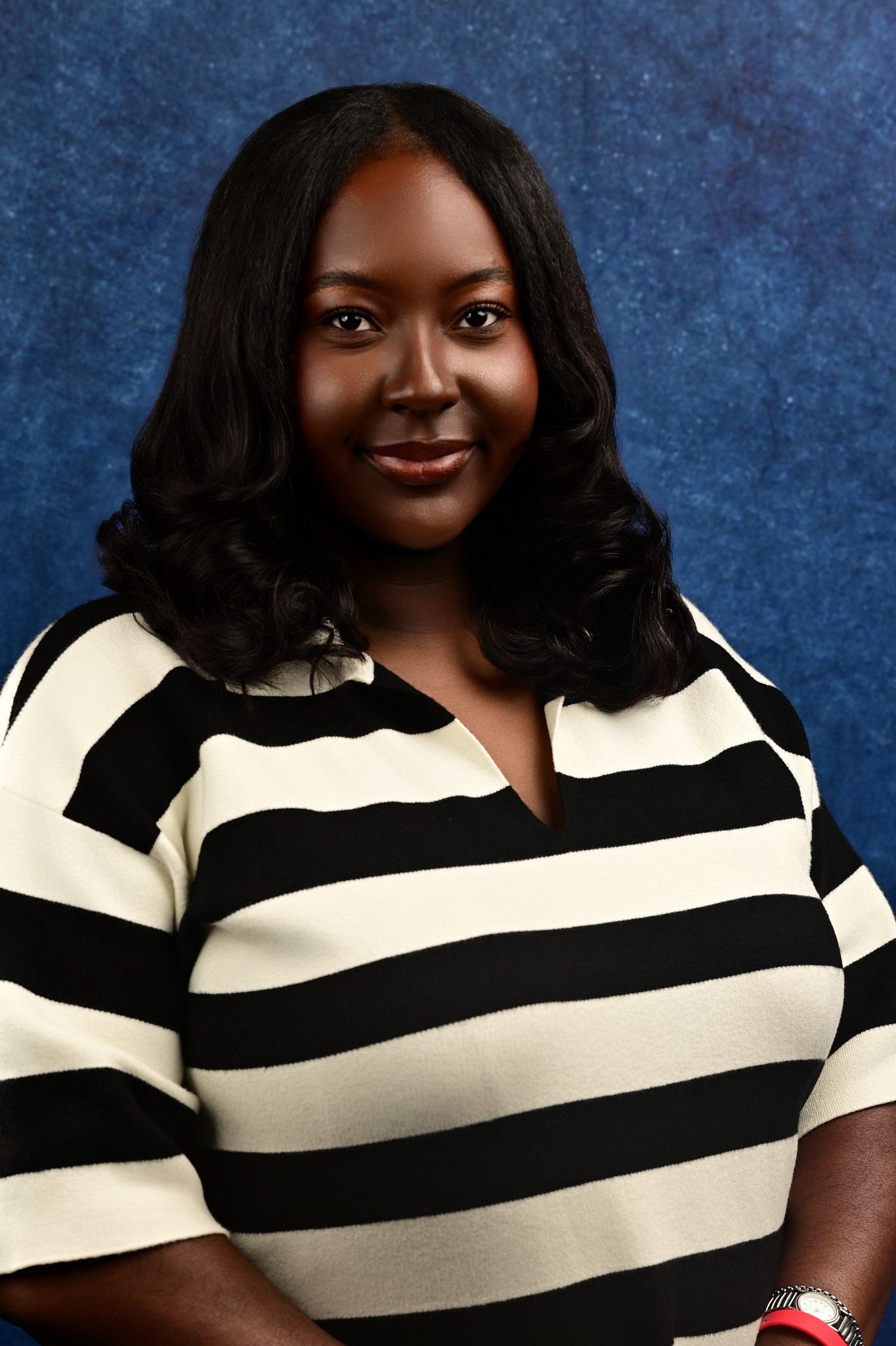 Smiling woman with shoulder-length wavy black hair wearing a black and cream striped top against a textured blue studio backdrop.