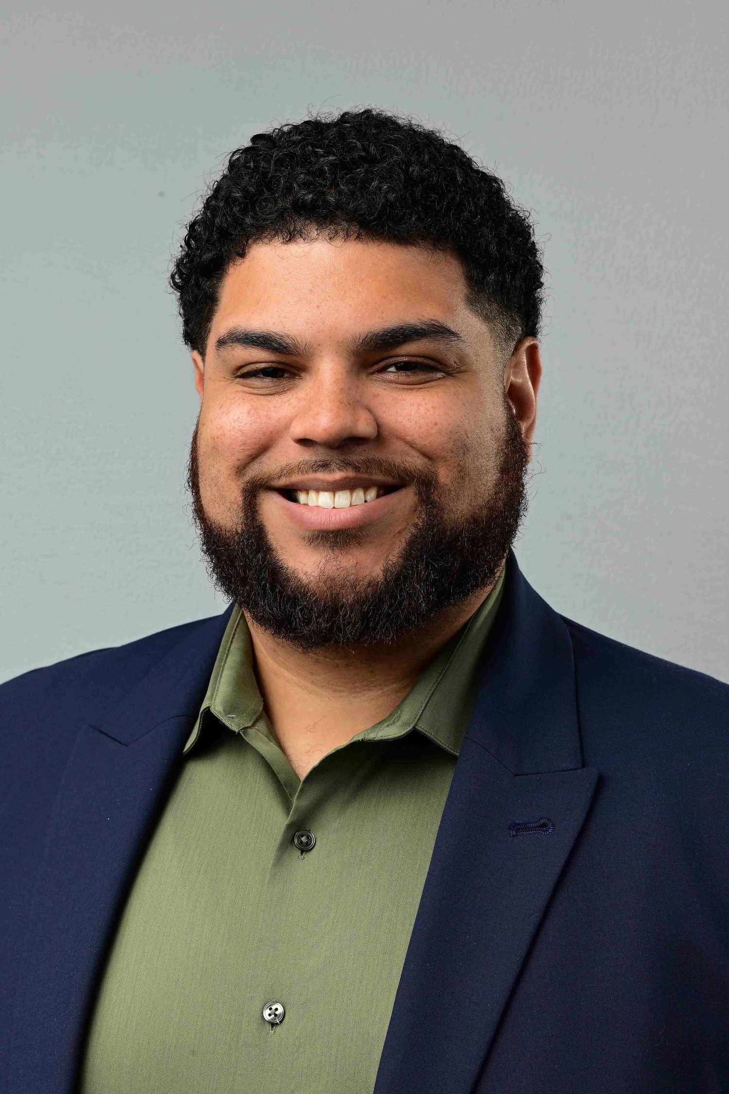 Smiling young man with beard wearing navy blazer and olive shirt against gray background.