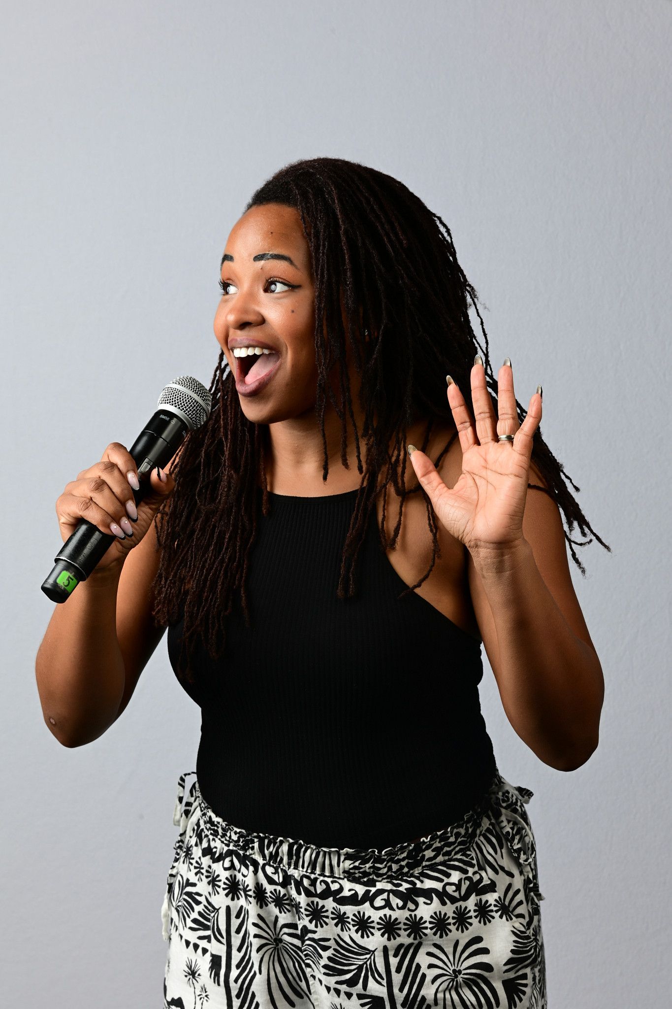 A woman with dreadlocks sings into a microphone, raising her hand, wearing a black top and floral skirt.