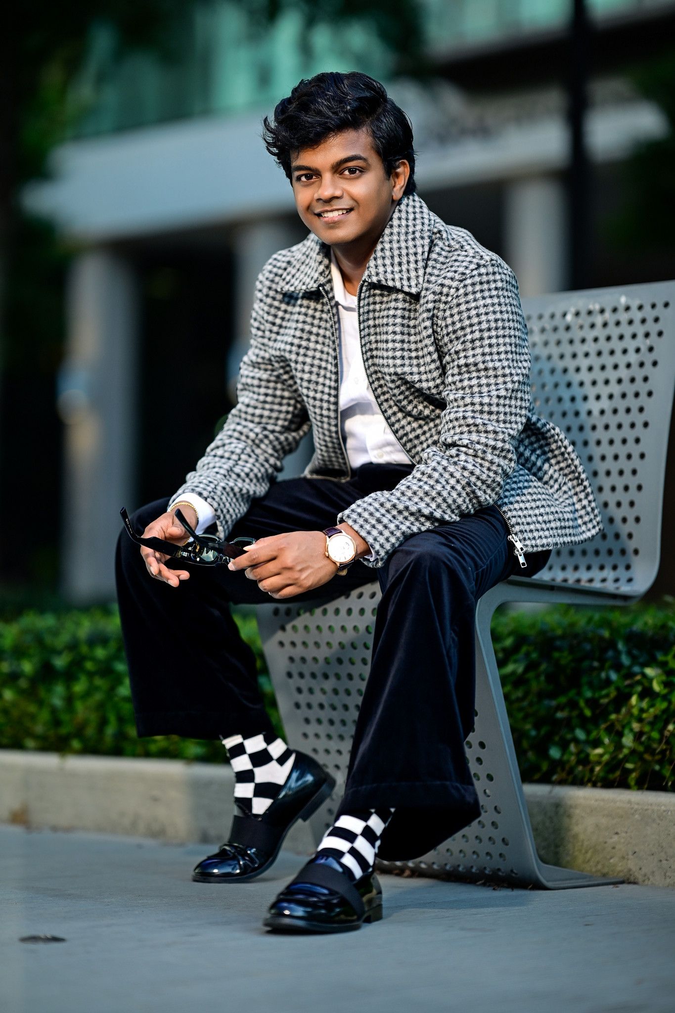 Young man in houndstooth jacket and checkered socks sits on bench holding camera, smiling outdoors.