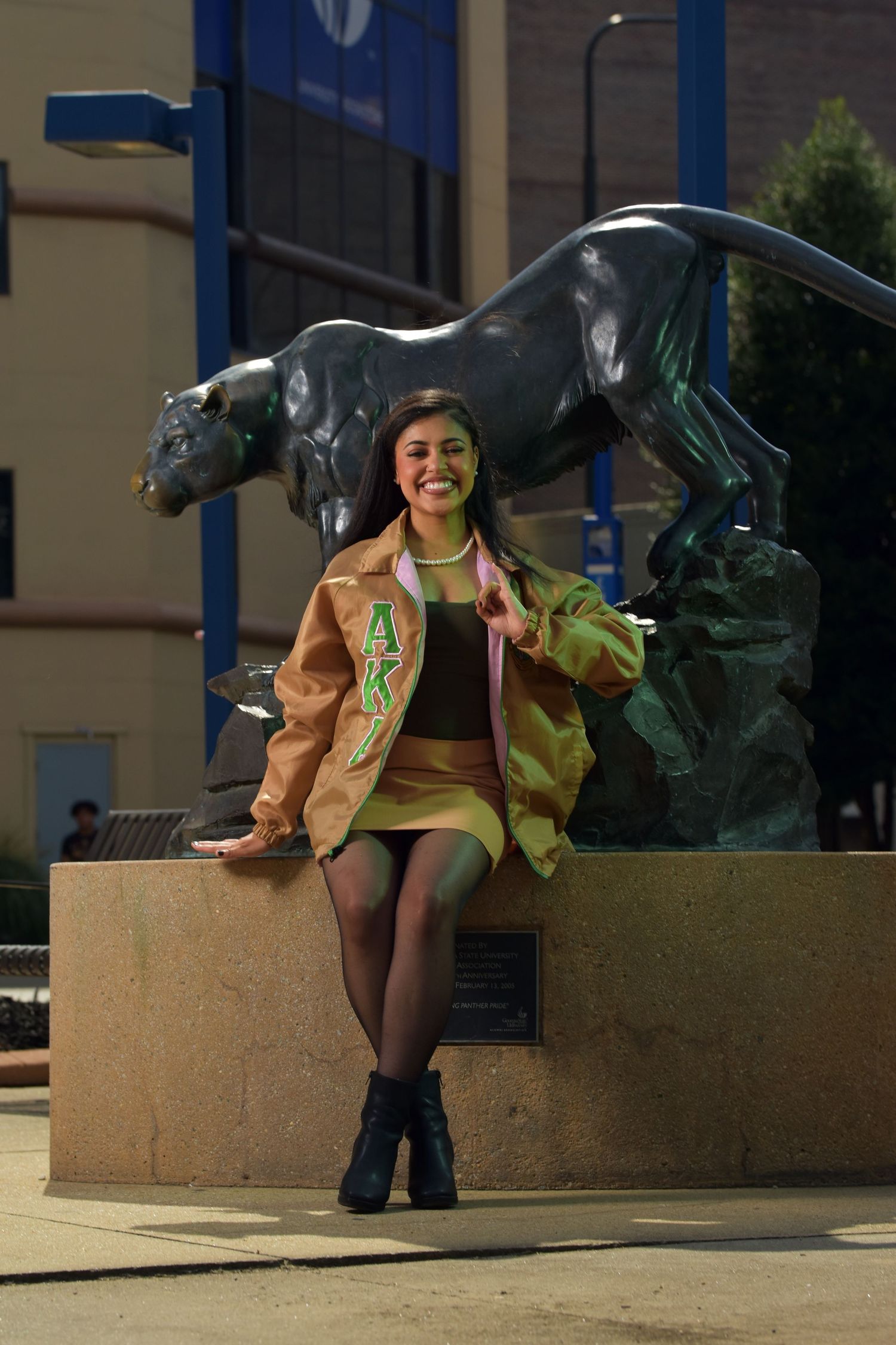 Smiling young woman in AKA sorority jacket poses beside panther statue on college campus.