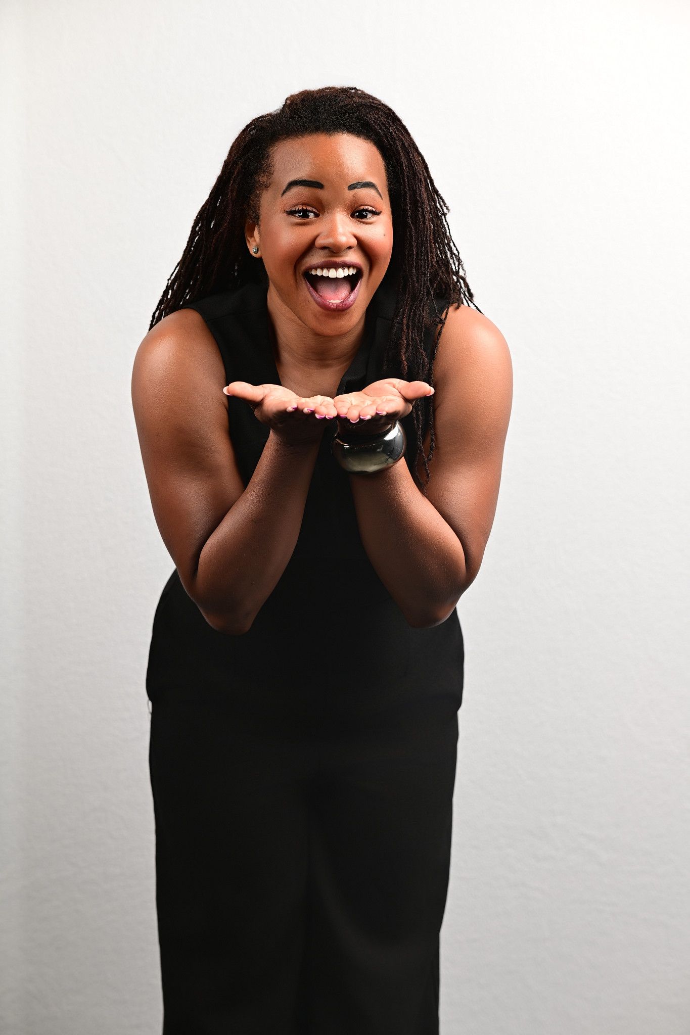 Woman with long locs in a black sleeveless dress leans forward with an excited open-mouthed smile and cupped open hands.