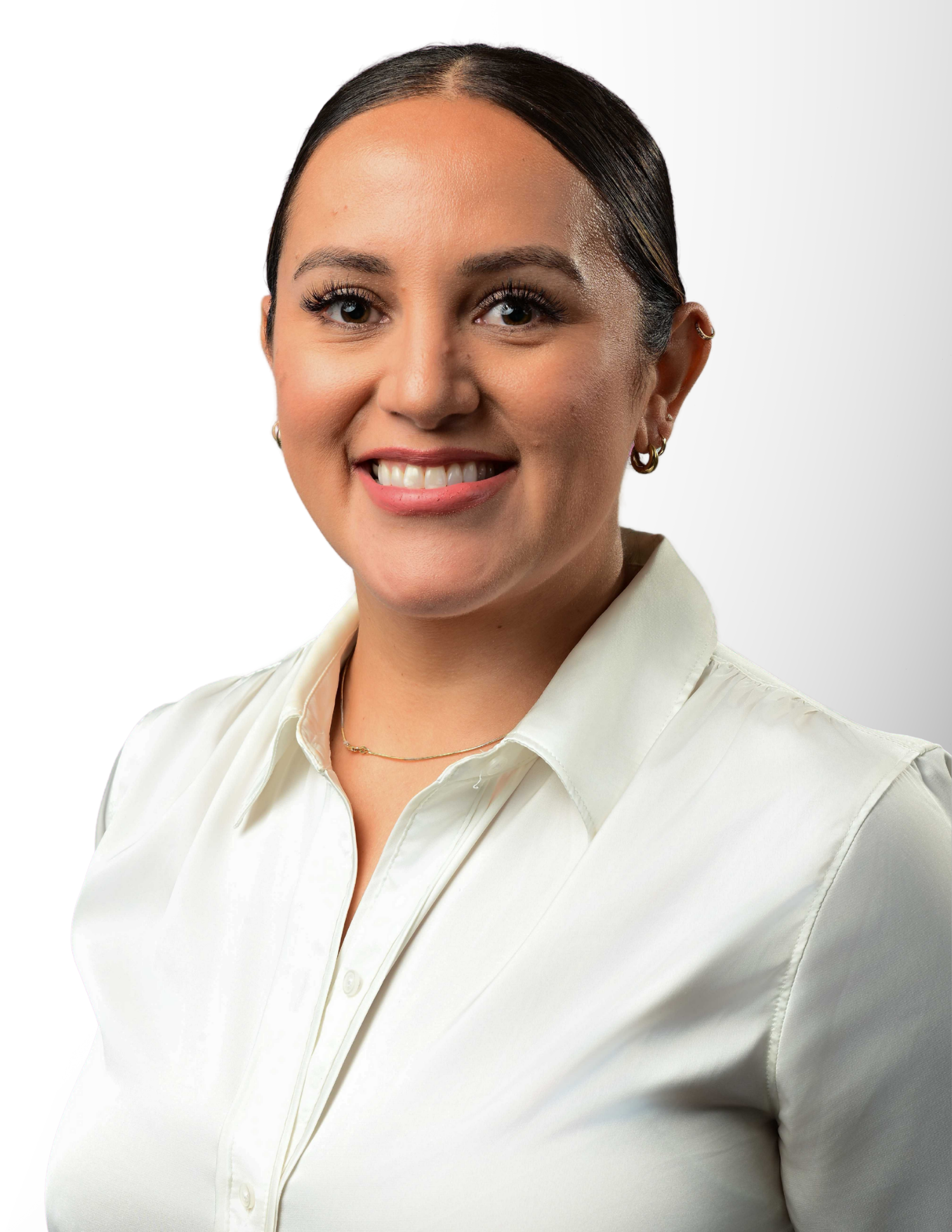 Professional headshot of a smiling young woman in a white sleeveless blouse with hair pulled back, wearing gold earrings and necklace.