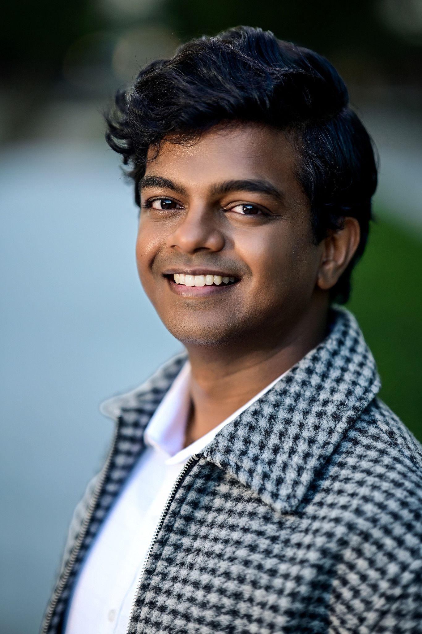 Young South Asian man smiling confidently outdoors, wearing a houndstooth blazer over a white shirt.