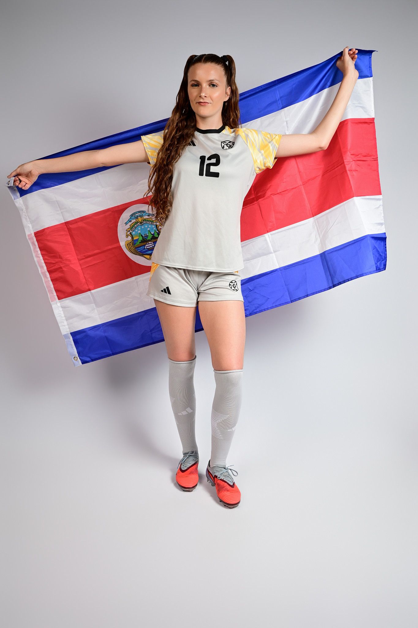 Female soccer player #12 in white Adidas kit holds Costa Rica flag against gray background.