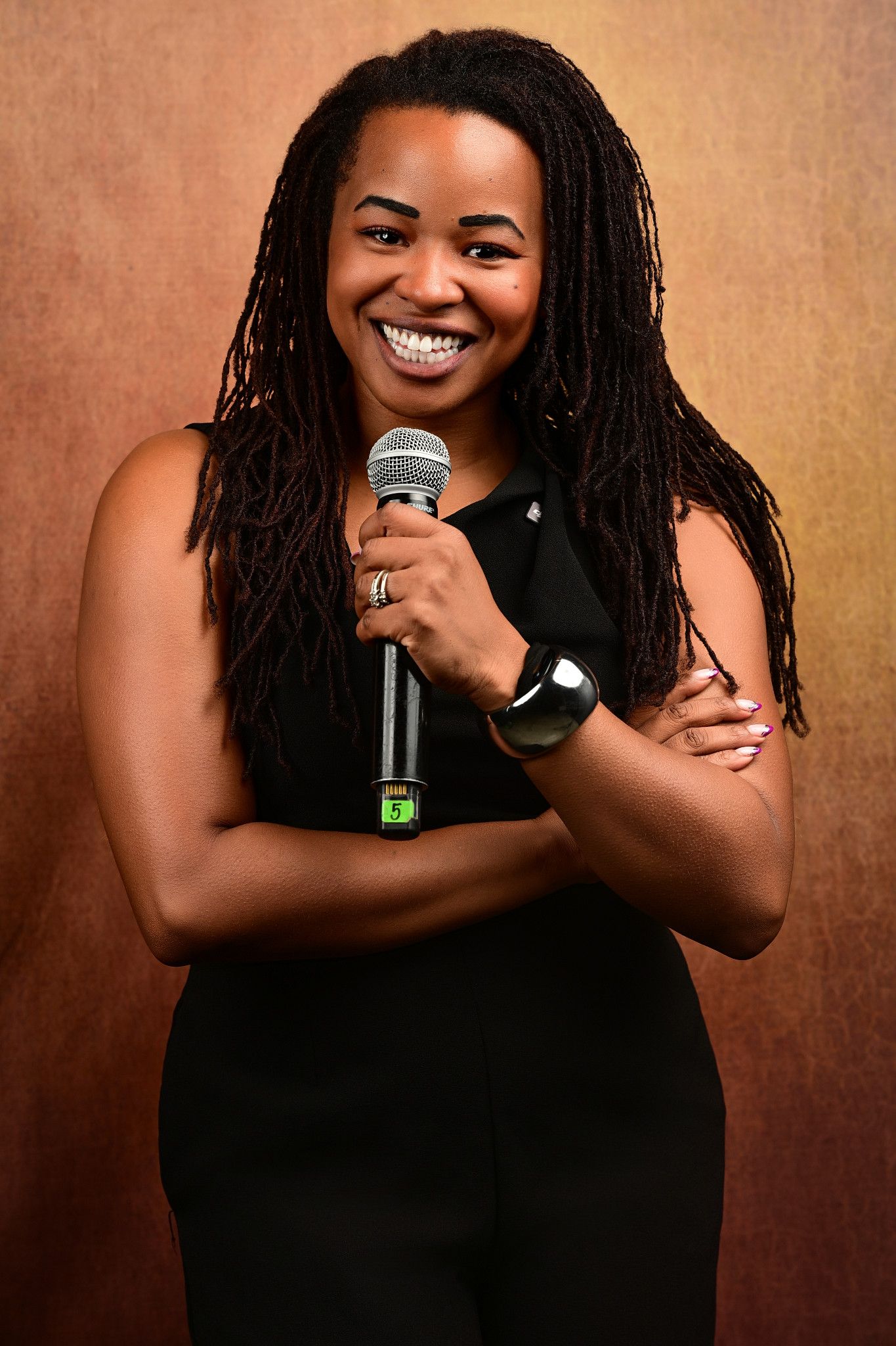 Smiling woman with long locs in a black sleeveless outfit holds a microphone with arms crossed against a warm gradient backdrop.