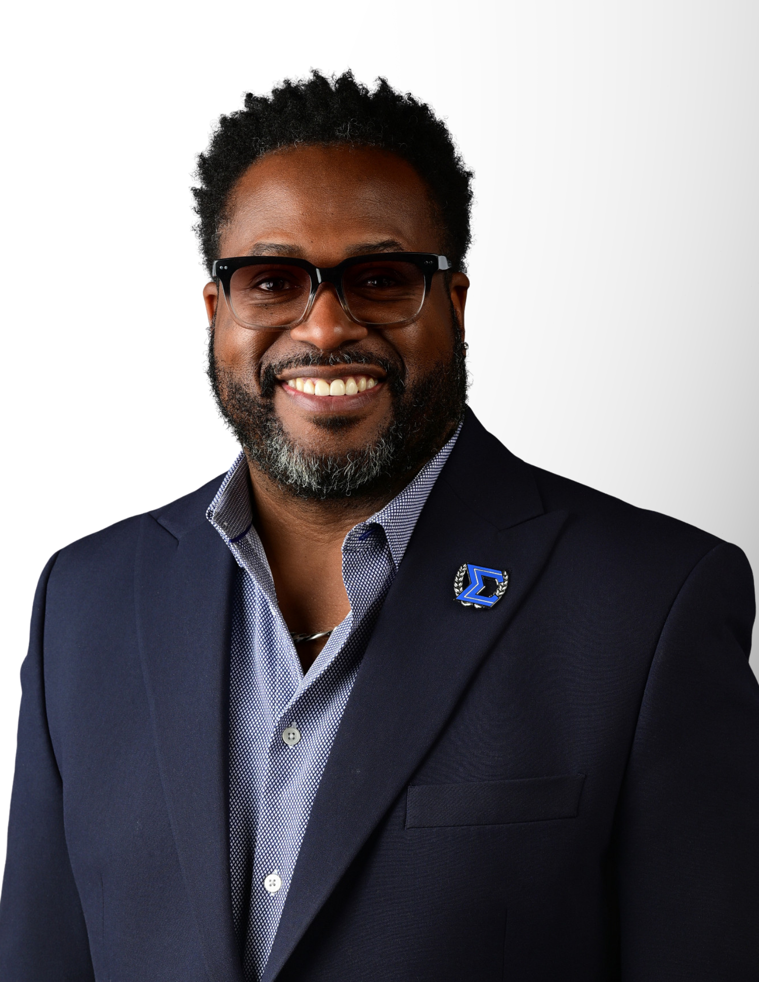 Professional headshot of a smiling man wearing a navy blazer, gray shirt, and brown sunglasses with a blue lapel pin.