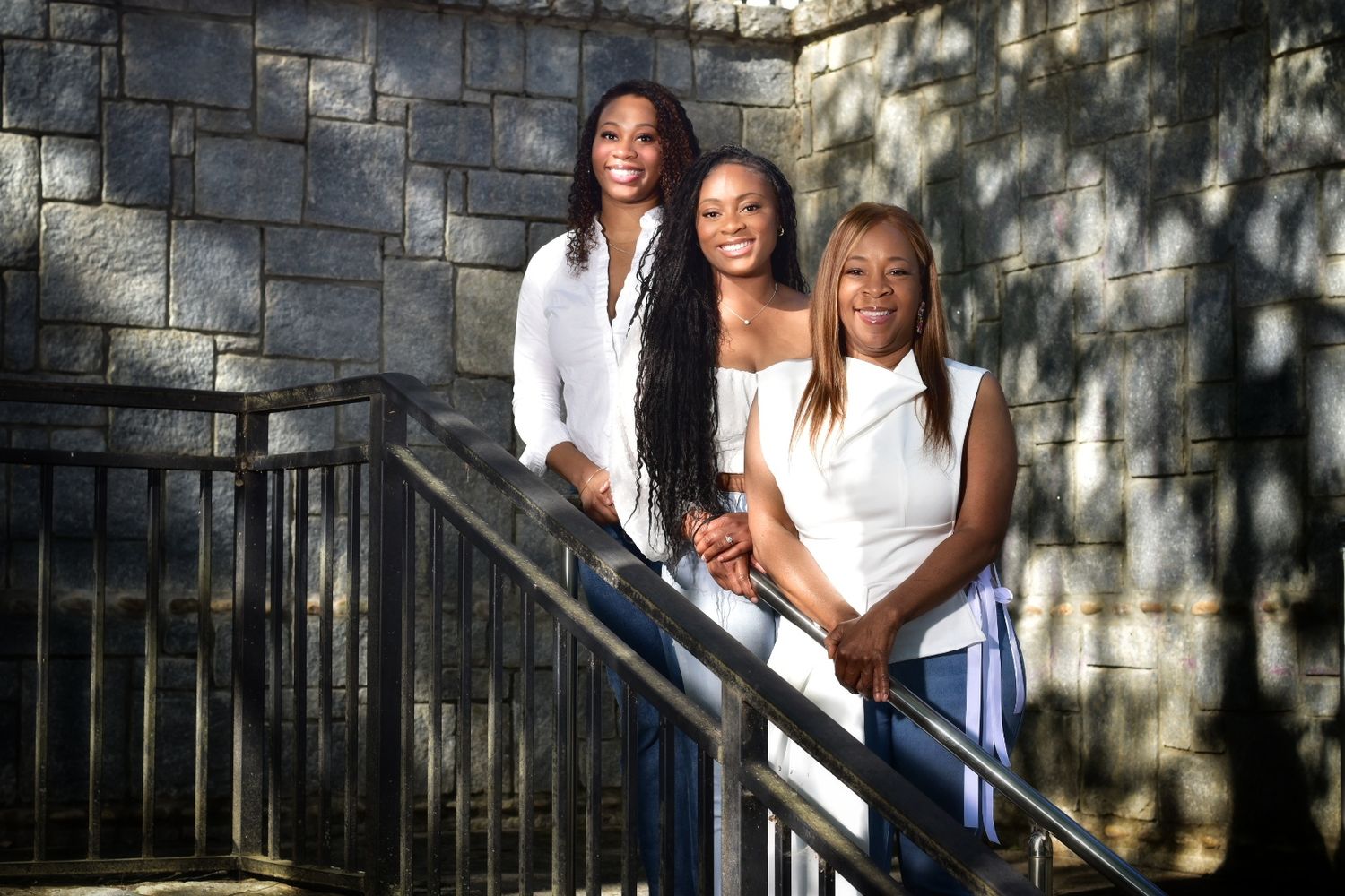 Three Black women in white tops pose together on outdoor stairs in front of a stone wall in sunlight.