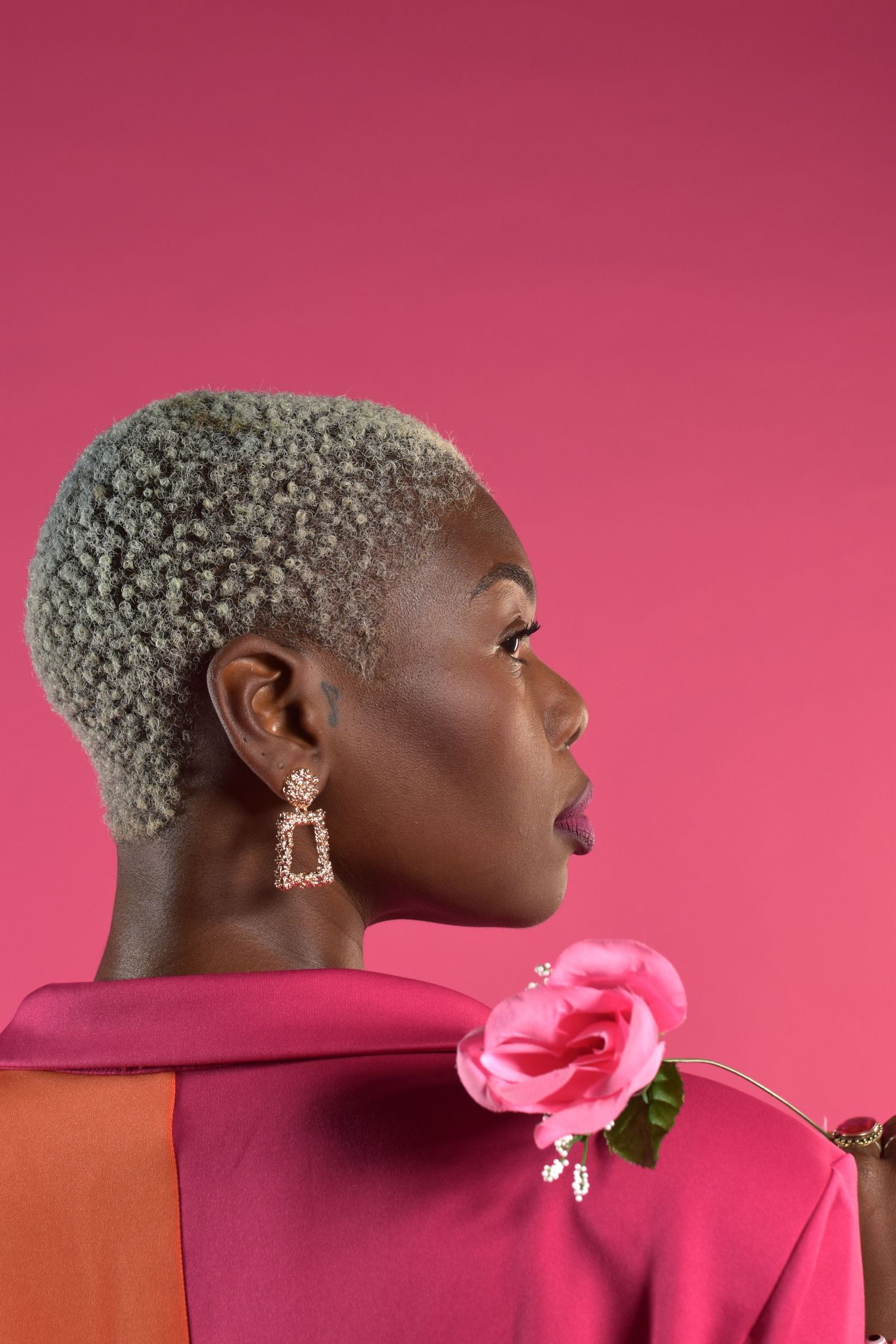 A Black woman with short gray hair wears a pink blazer with a rose, viewed from behind in profile.