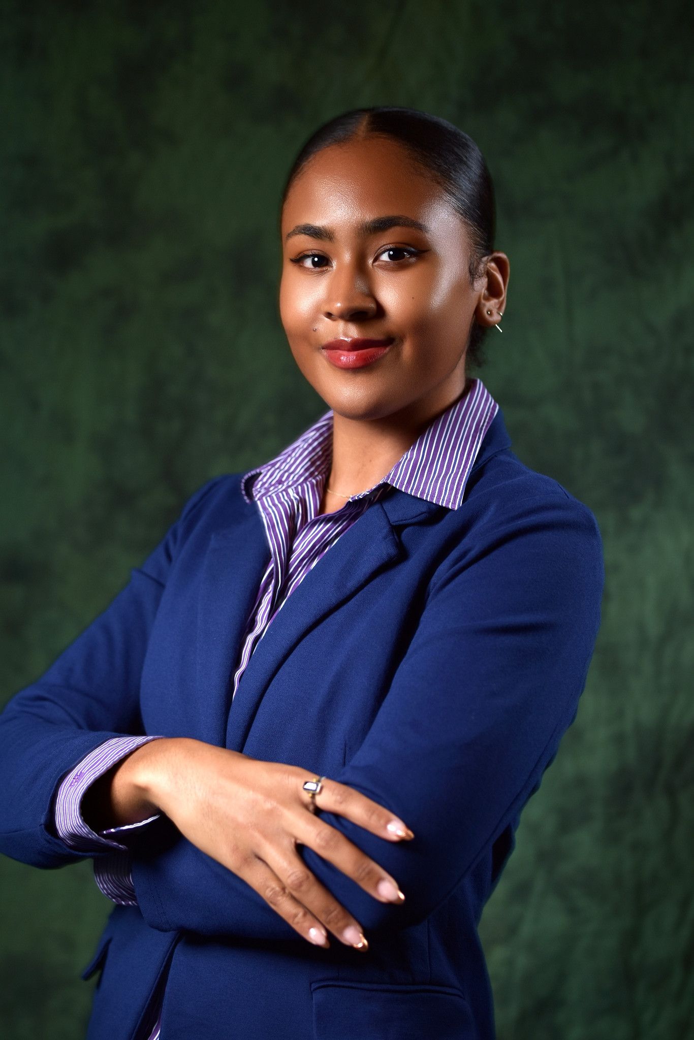 Professional woman in blue blazer and striped shirt stands with arms crossed against dark green background.